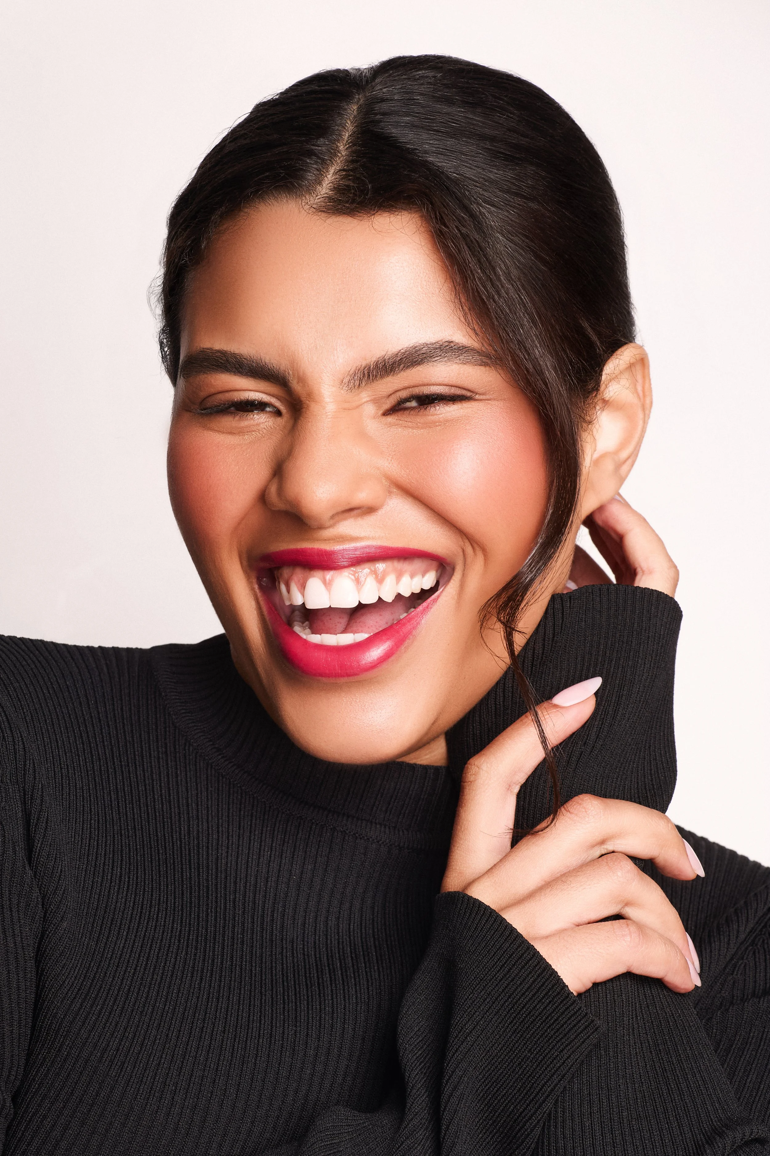 A woman laughing with her mouth open, wearing a black turtleneck, with dark hair pulled back.