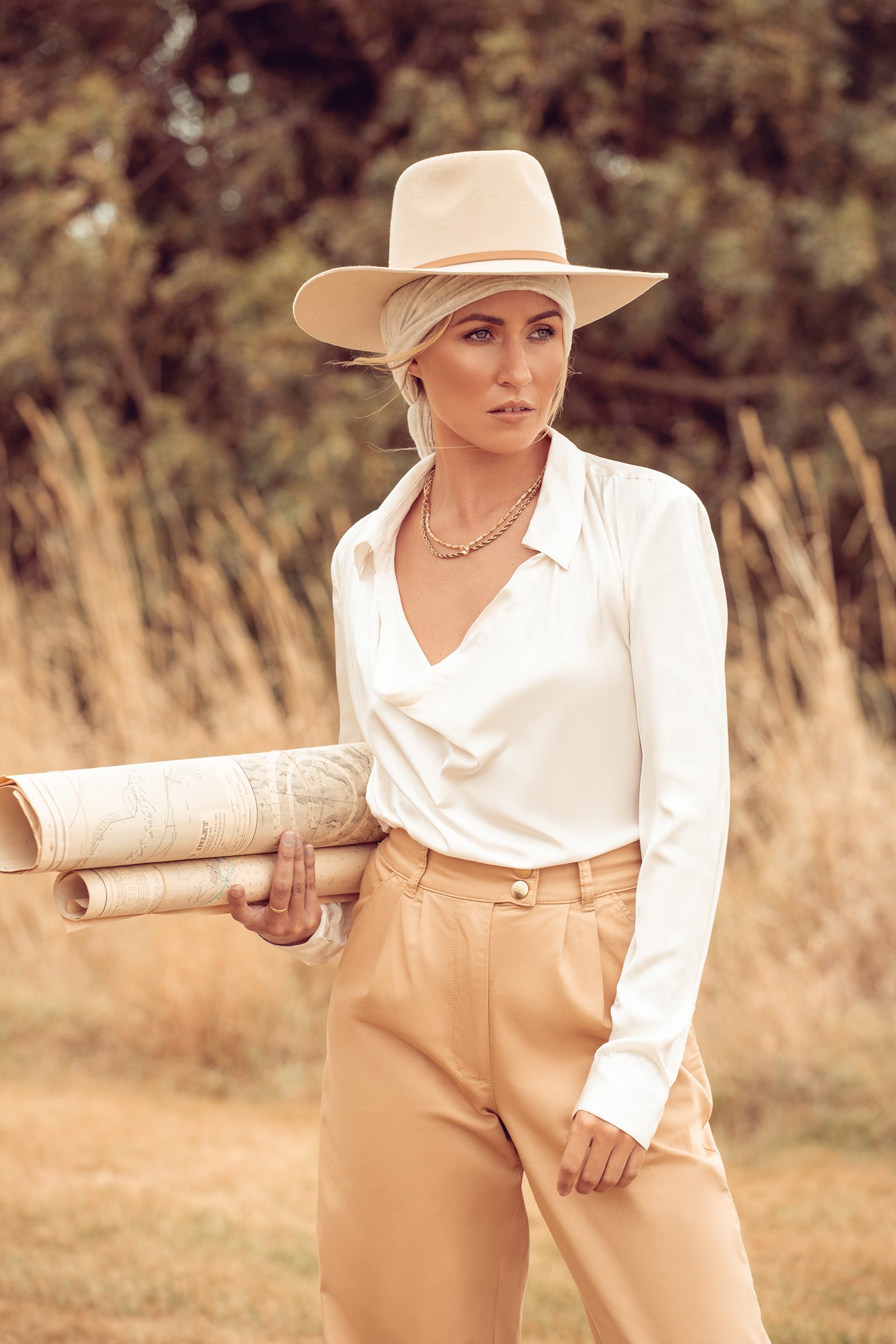 Woman in a white blouse and beige hat holding rolled maps outdoors.