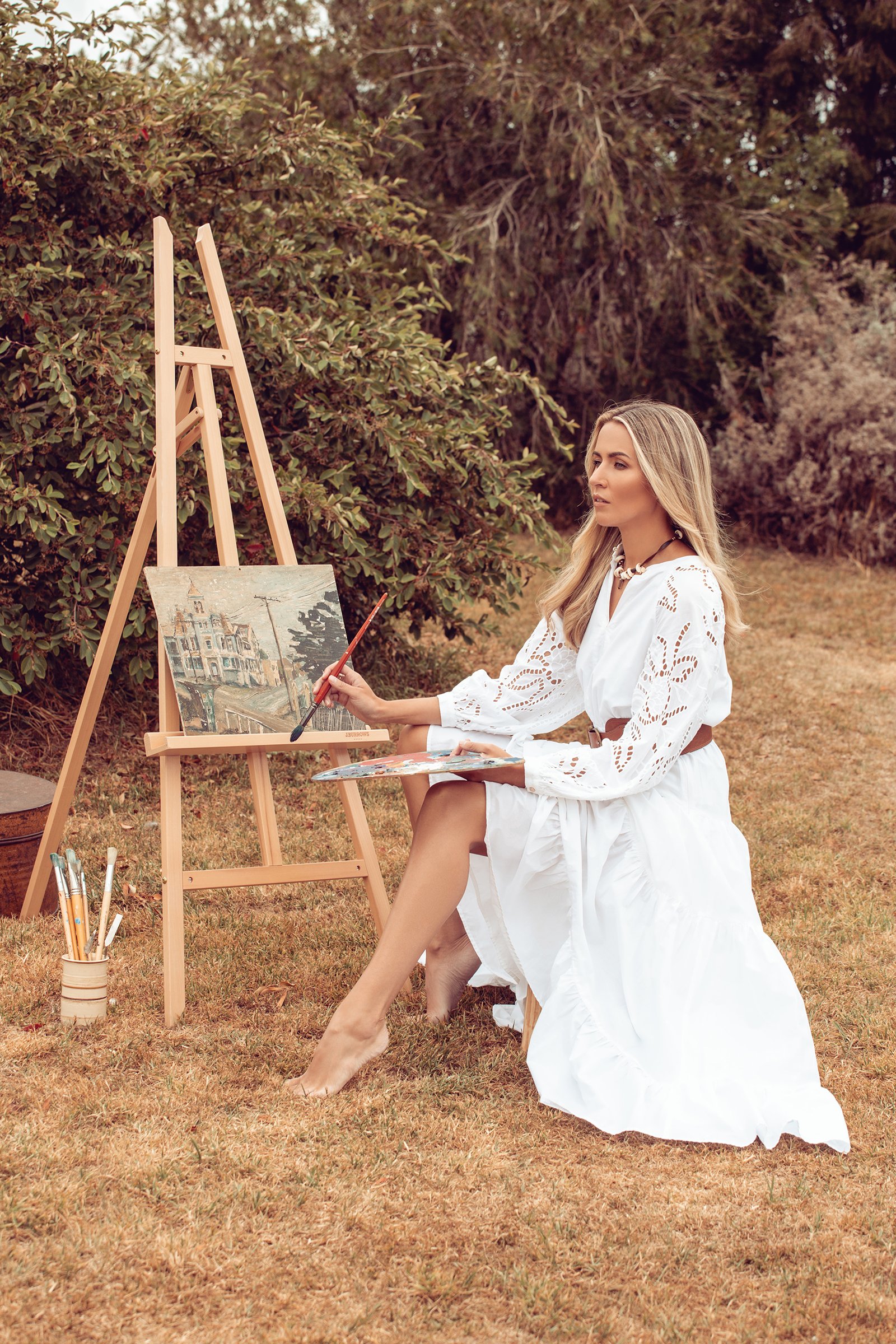 Woman in white dress painting on canvas outdoors with easel and brushes, surrounded by greenery.