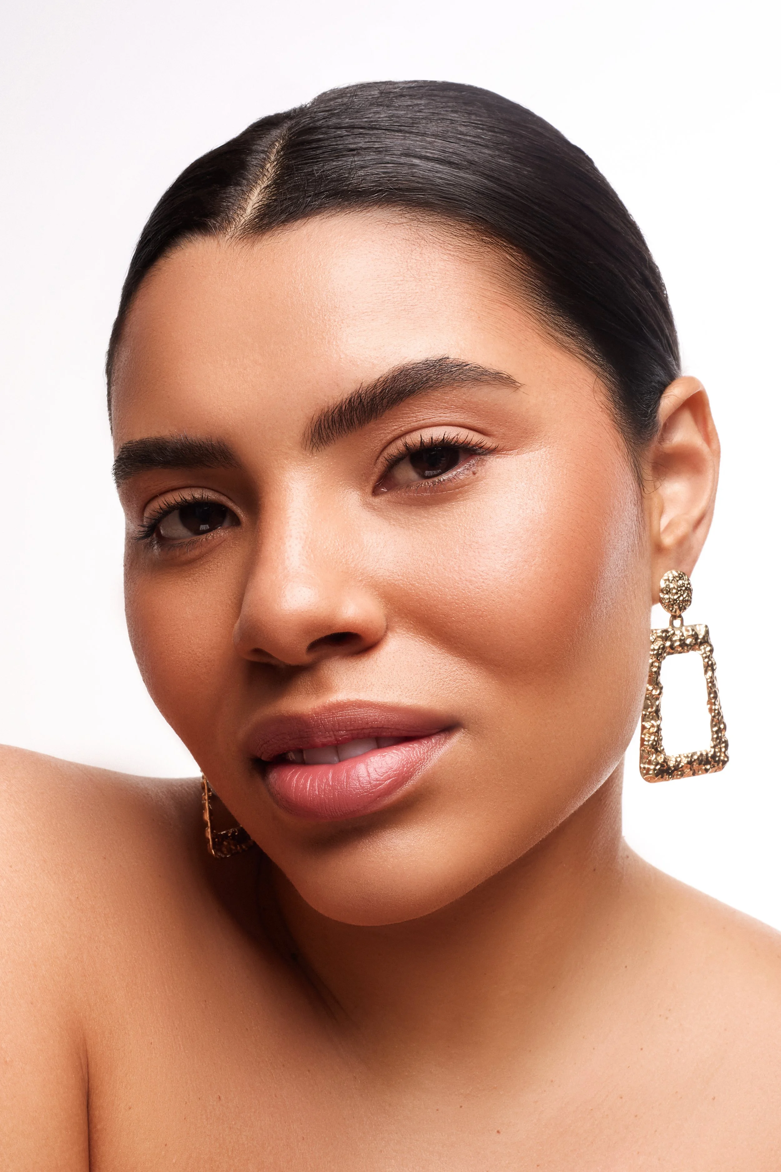 A close-up portrait of a woman with smooth skin, dark hair styled back, and wearing large, gold square-shaped earrings.