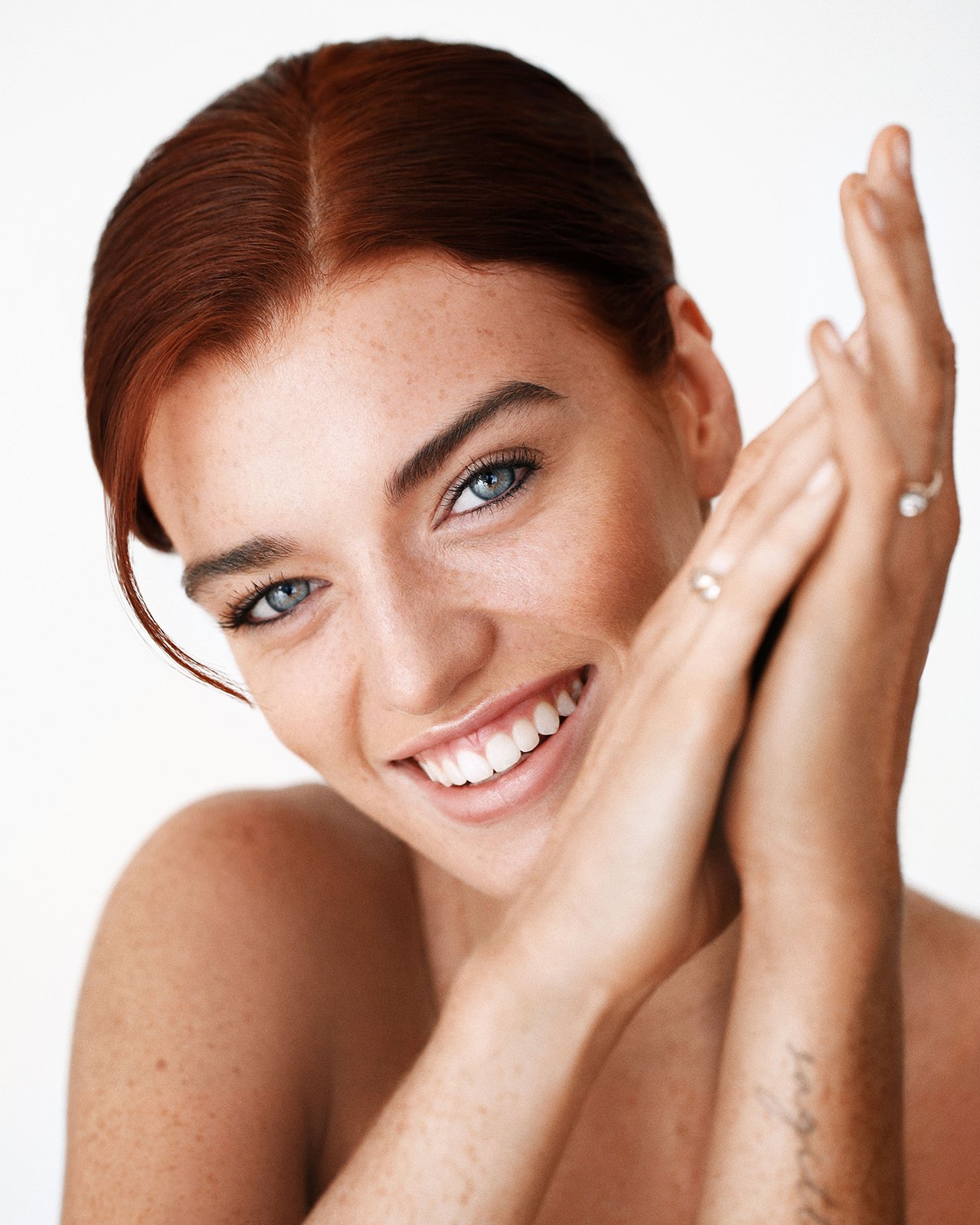 Smiling person with red hair and blue eyes, clasping hands in front of face.