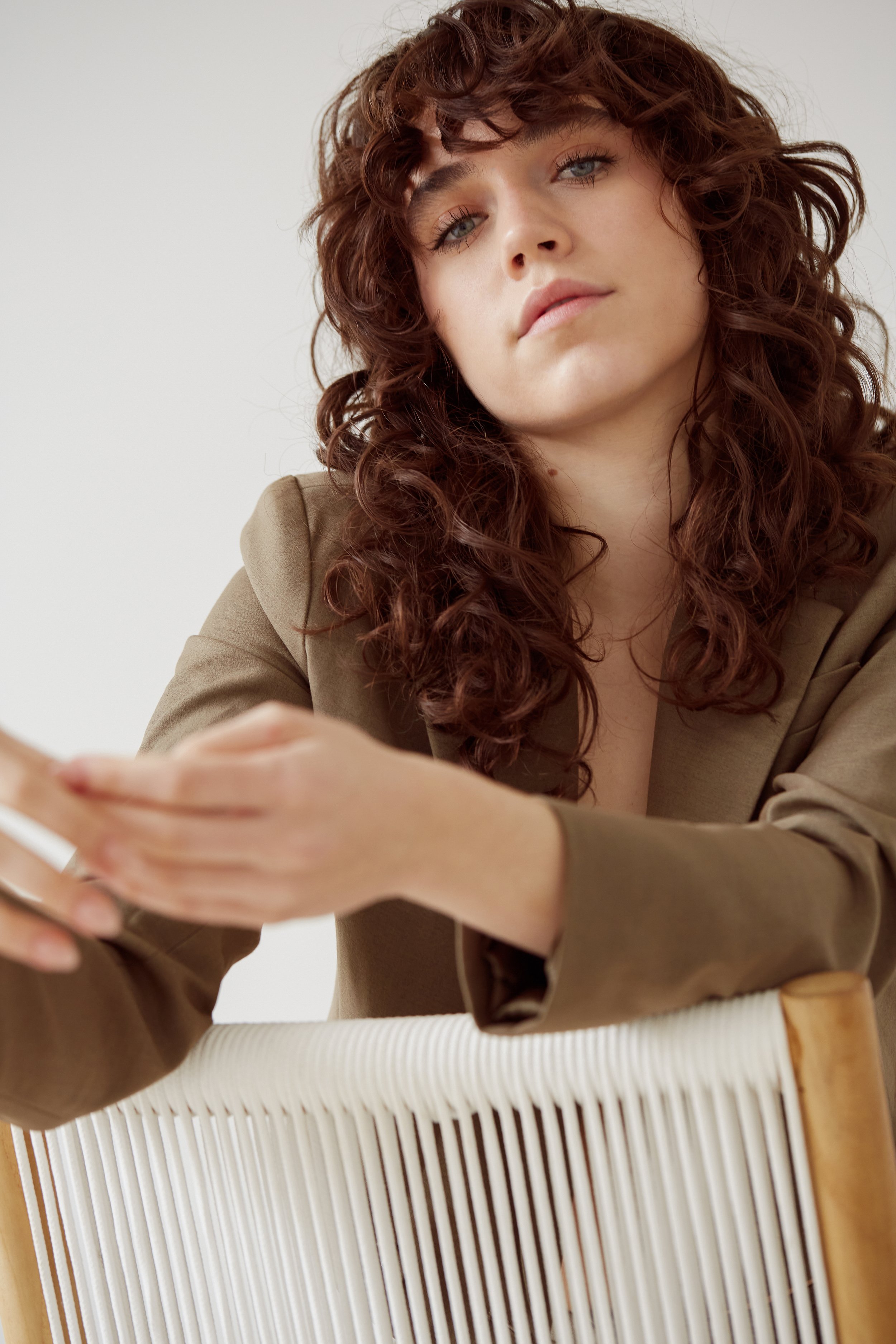 Person with curly hair sitting on a chair, wearing a brown jacket, looking at the camera, neutral background.