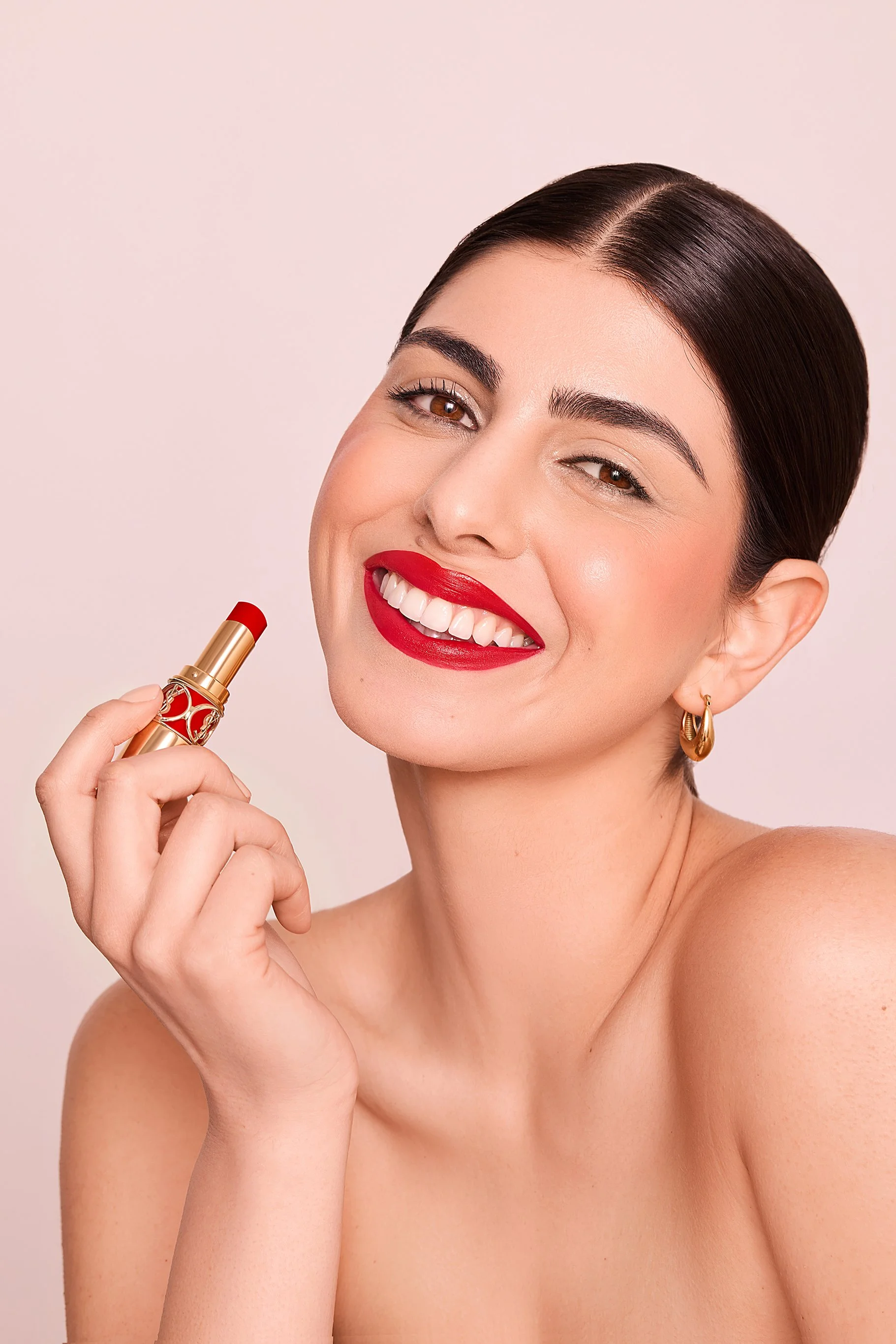 Smiling woman holding a red lipstick near her lips, wearing red lipstick and gold hoop earrings.