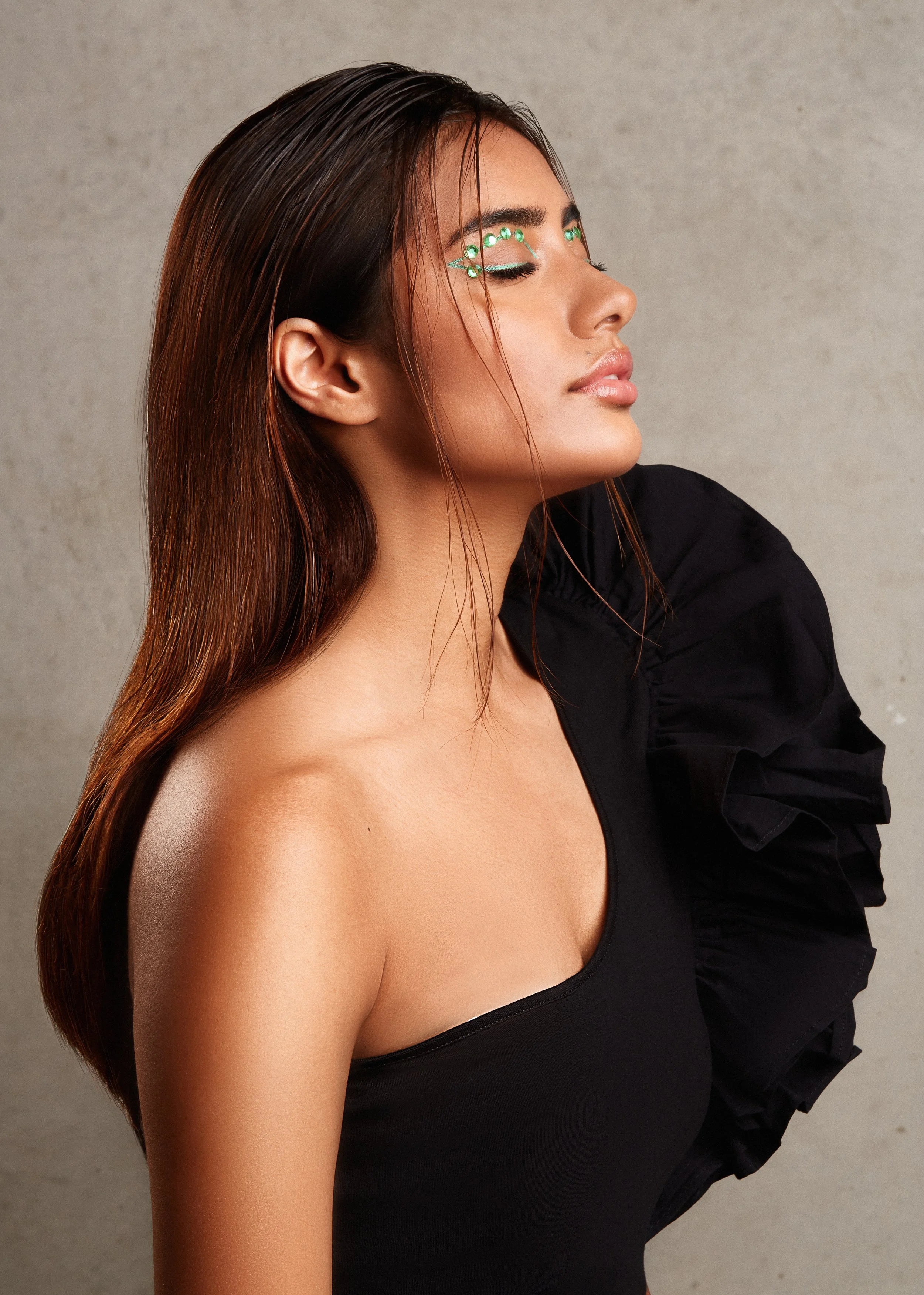 Profile of a woman wearing a black one-shoulder top with decorative shoulder ruffle, eyes closed, adorned with green facial gems, against a gray background.