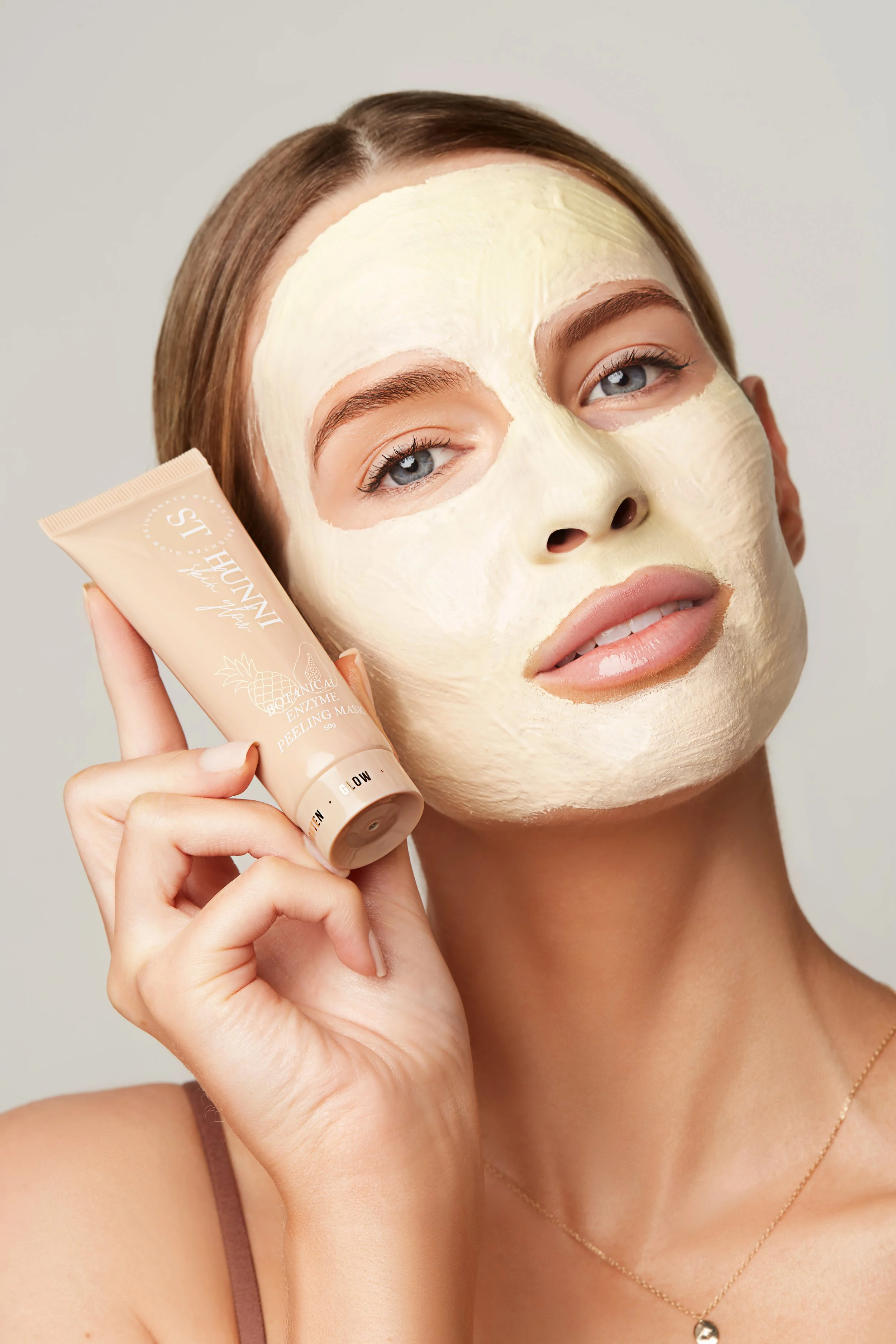 Person with a facial mask holding a St. Hunni Botanical Enzyme Peeling Mask tube.
