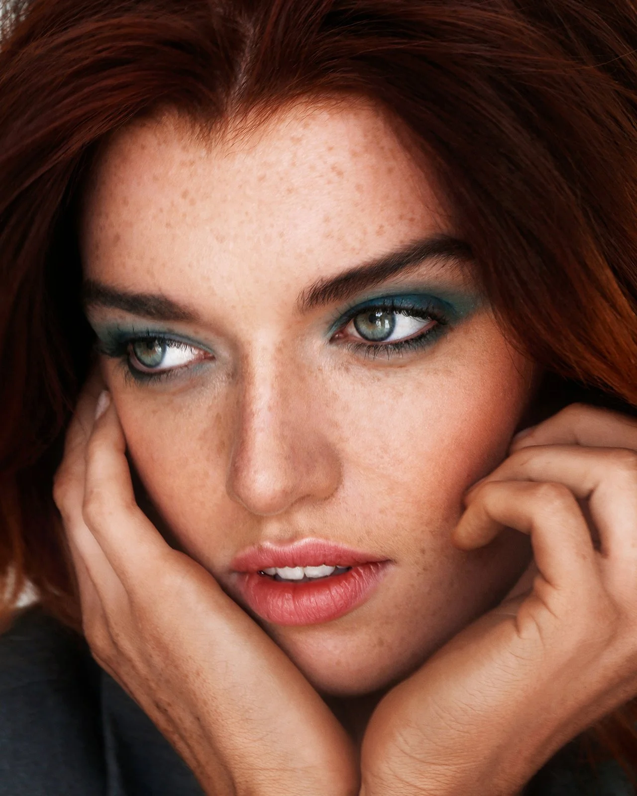 Close-up of a woman with red hair and blue eyeshadow, holding her face with her hands.