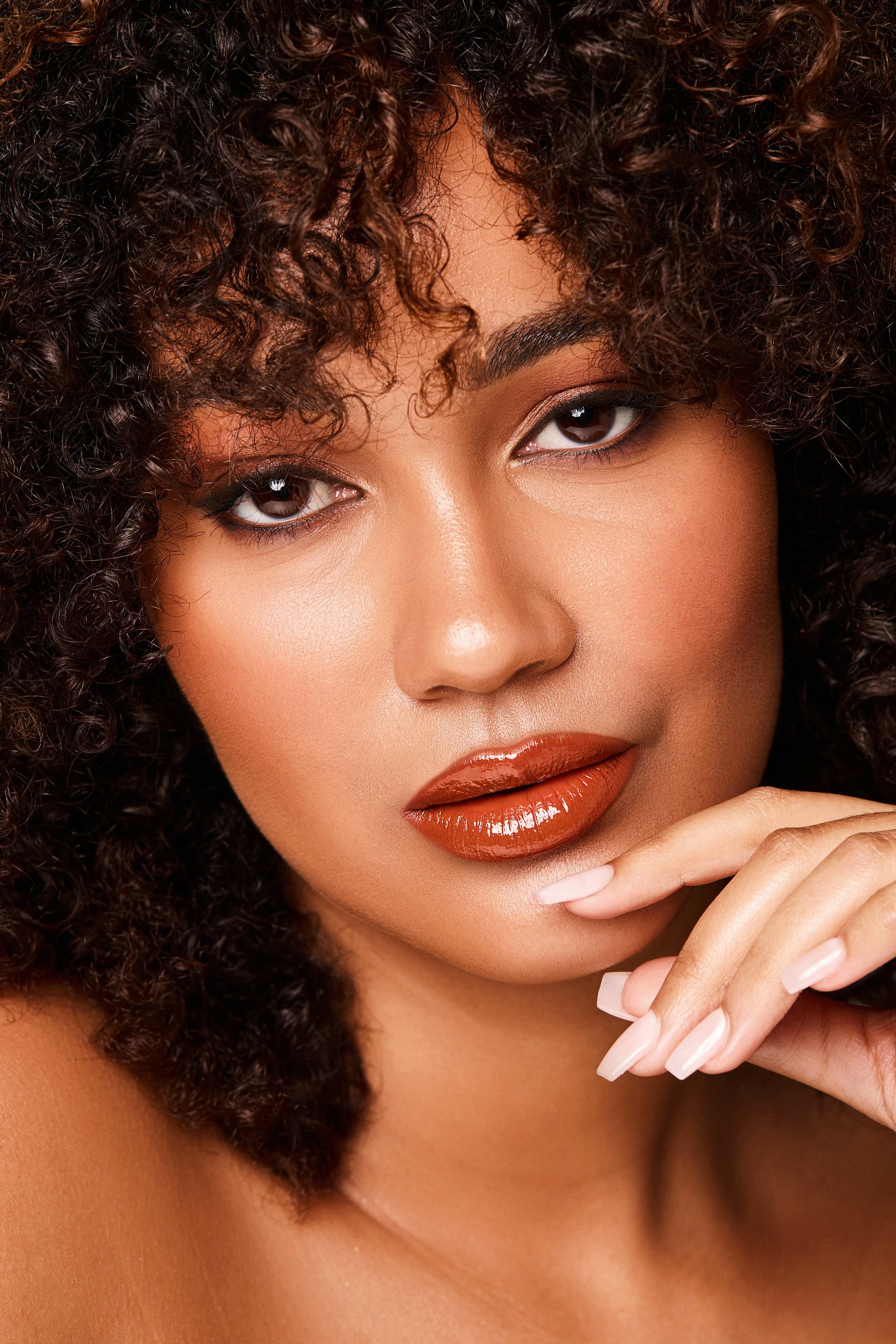 Close-up of a woman's face with curly hair, makeup, and glossy lips, gently touching her chin.