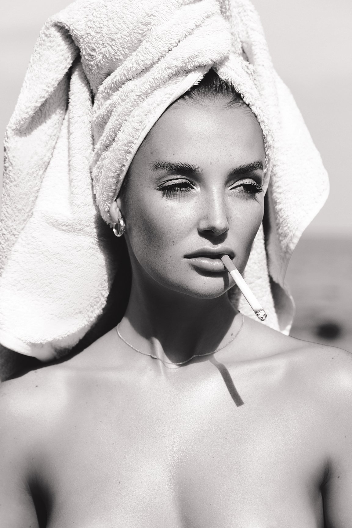 A woman with a towel wrapped around her head, holding a cigarette in her mouth, in a black and white photograph.