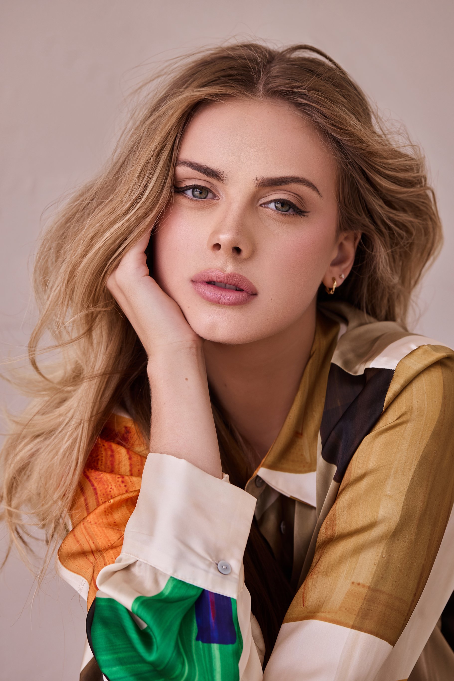 Woman with long blonde hair in colorful shirt posing, head resting on hand.