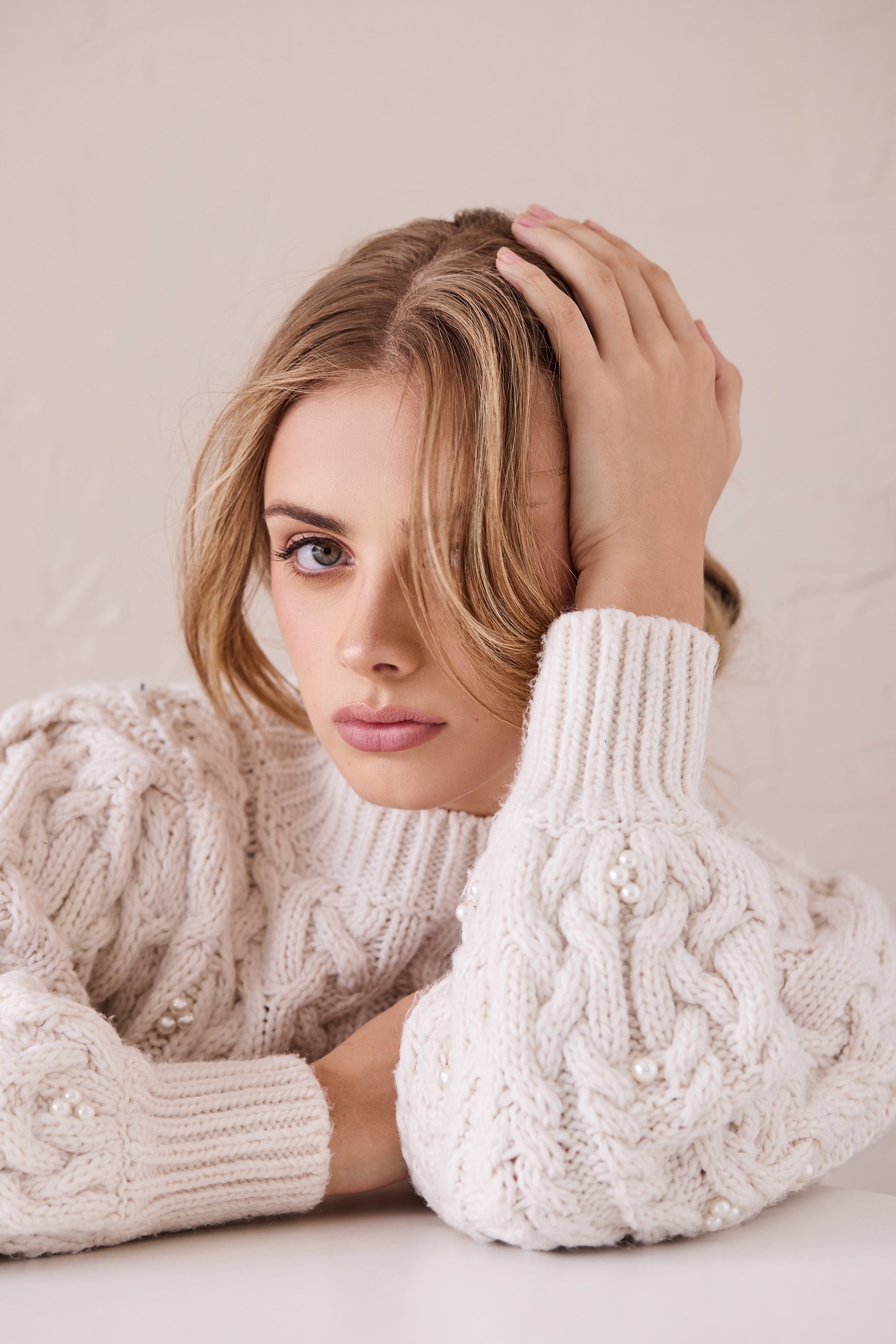 Person in a white cable knit sweater with pearls, resting their head on their hand.