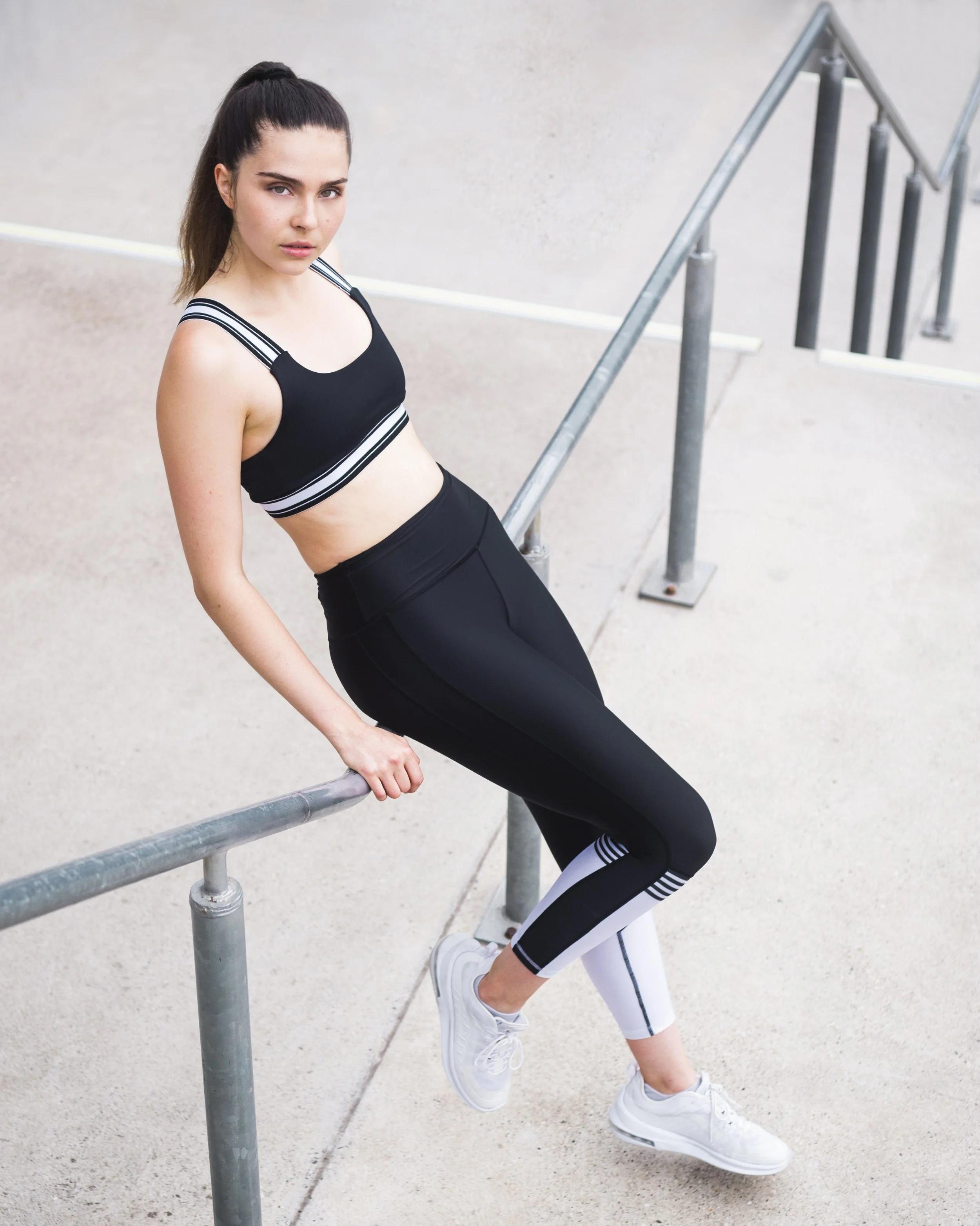 A woman wearing athletic wear, including a black sports bra and leggings, leaning on a metal railing outdoors, with white sneakers.