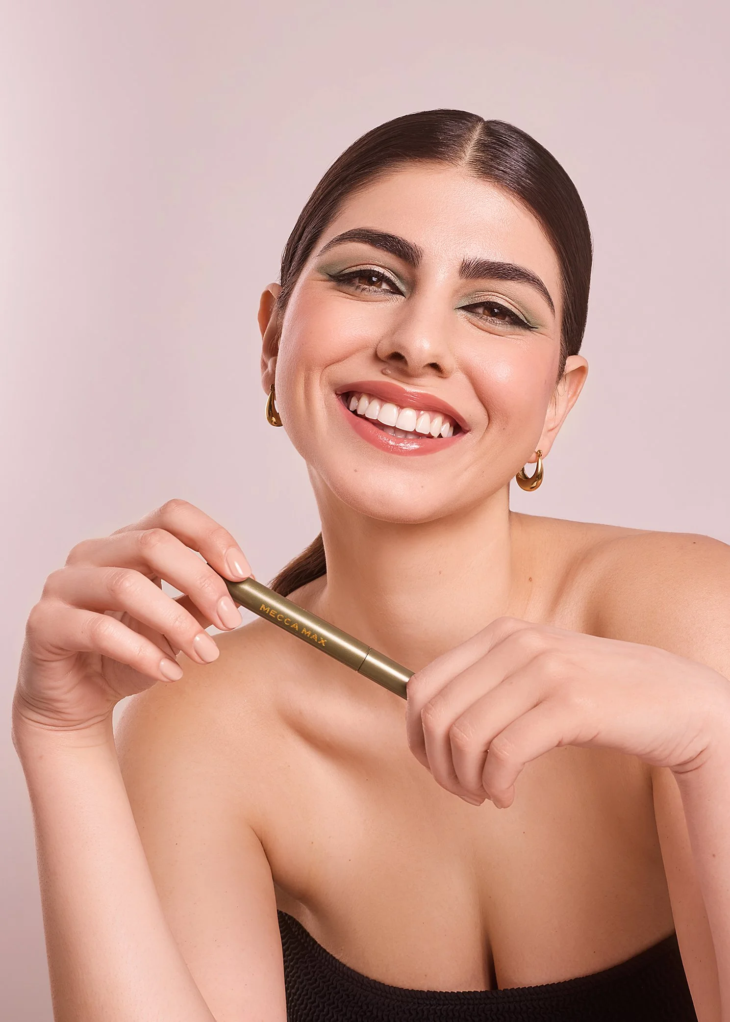 Smiling woman holding a Mecca Max mascara tube with bold eye makeup.