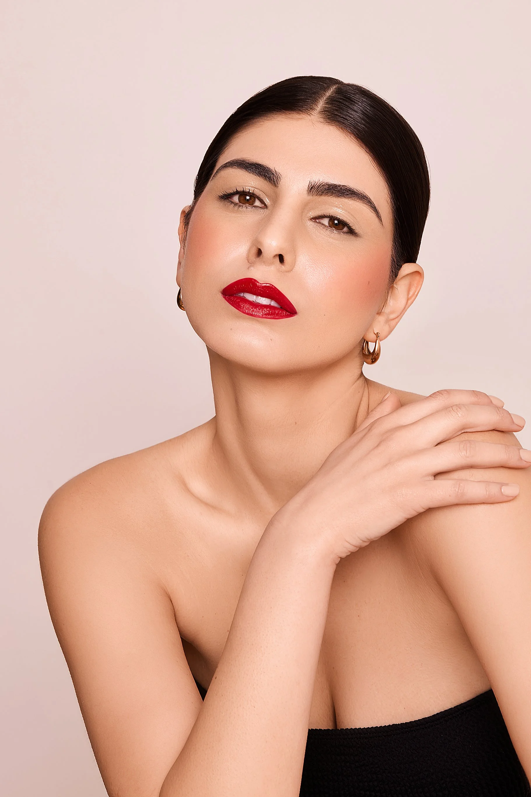 Portrait of a woman with dark hair, red lipstick, wearing hoop earrings and a black top against a beige background.