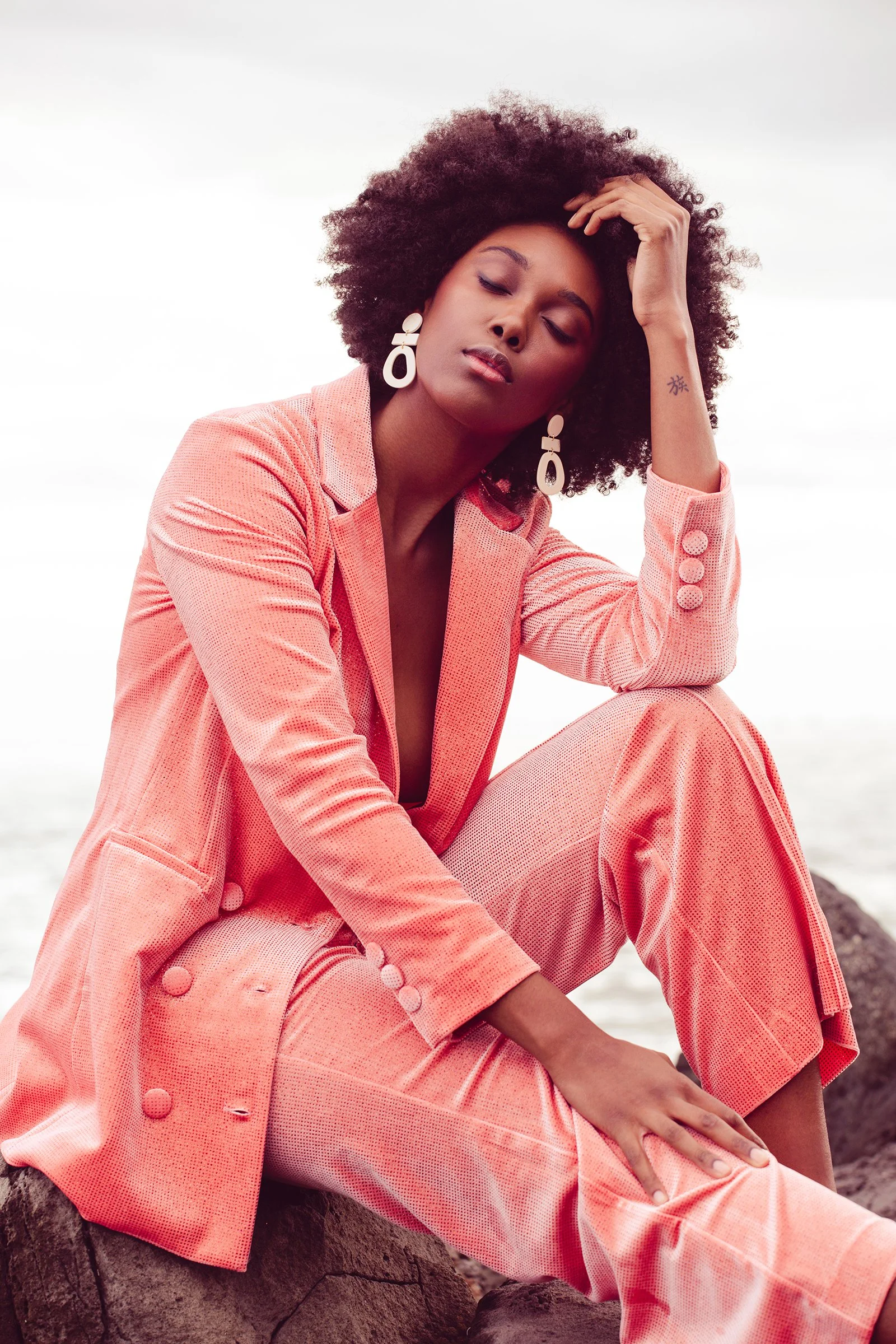 Fashion model wearing a coral pink suit with large buttons and white geometric earrings, sitting outdoors with eyes closed, hand on head, and a serene expression.