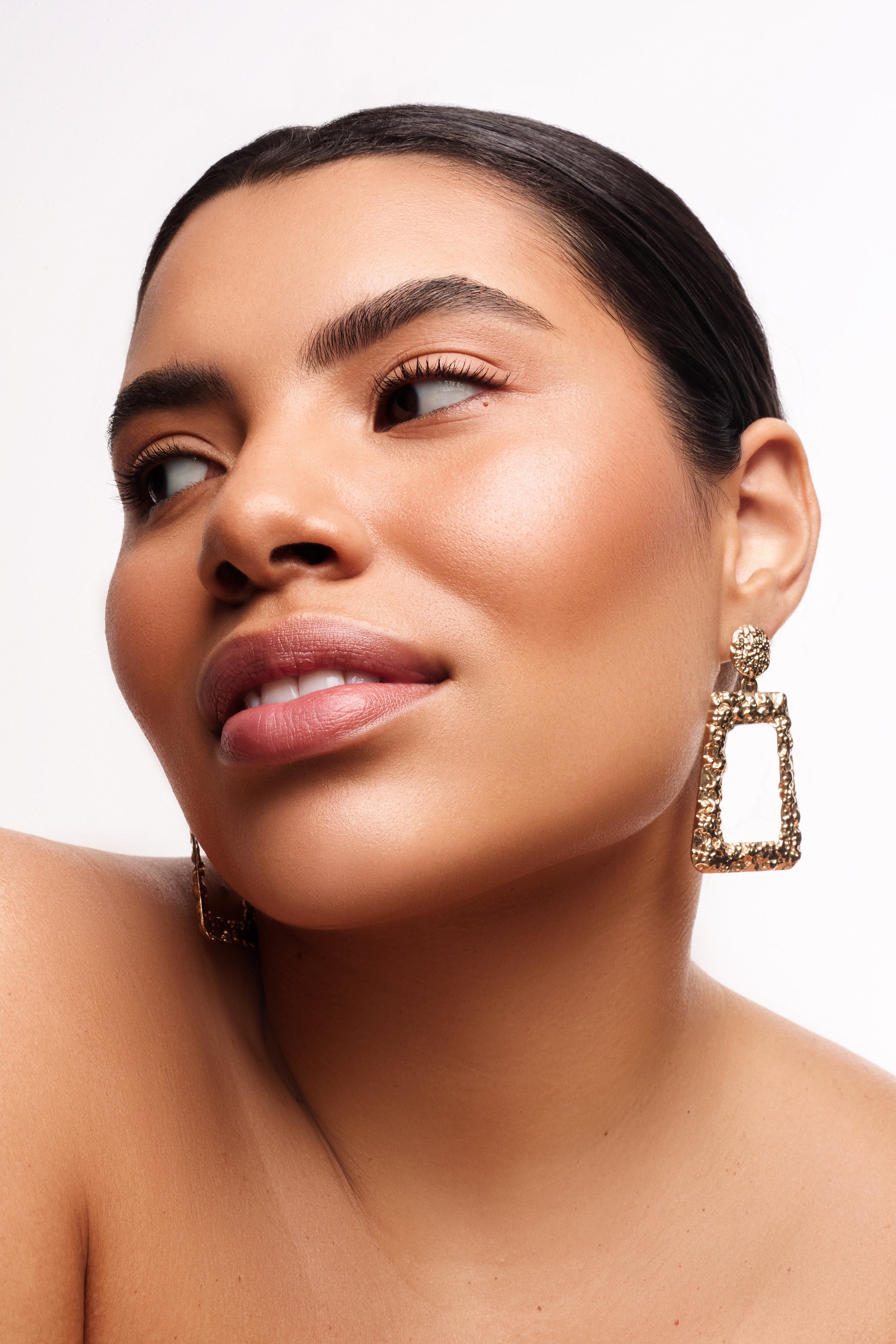 Close-up portrait of a woman with flawless skin, wearing gold earrings, with dark hair slicked back, looking slightly to her right with a subtle smile.