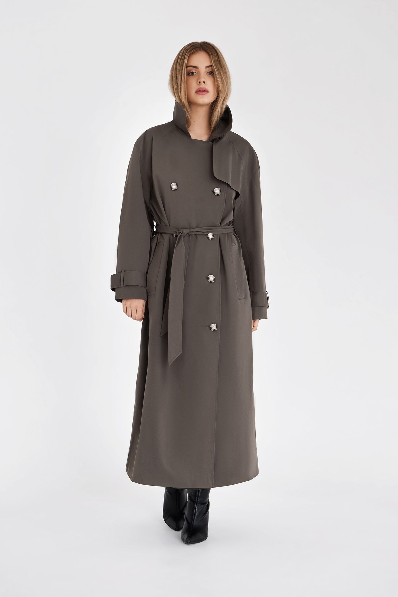 Person wearing a long gray coat with a belted waist and button details, standing against a plain background.