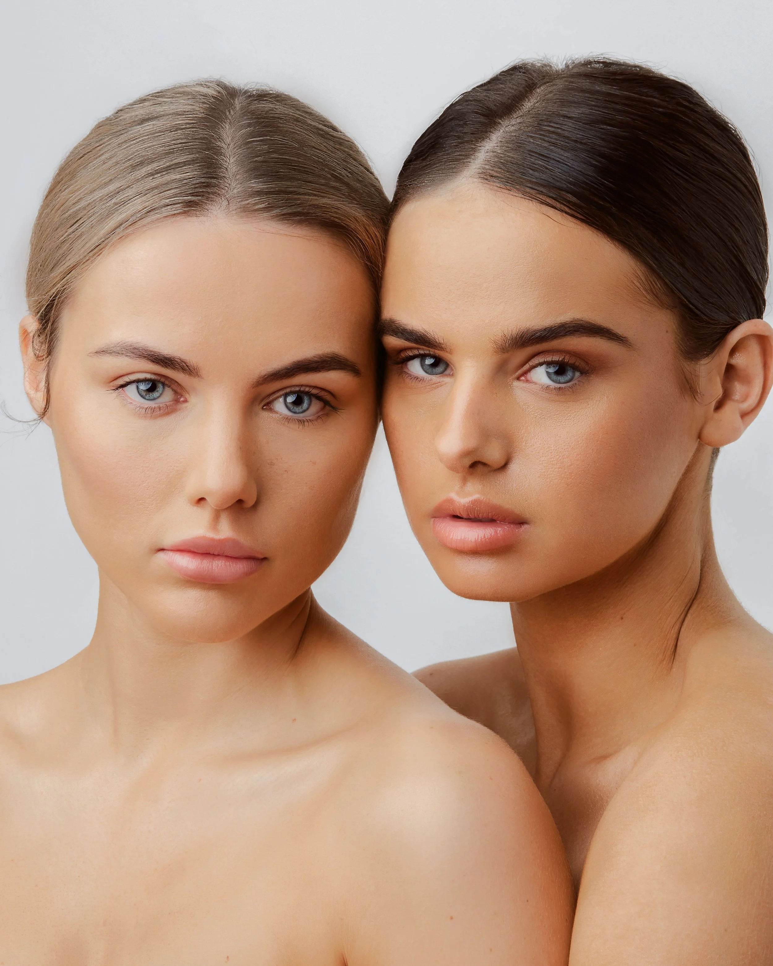 Two women with light makeup and neutral expressions, close together against a plain background.