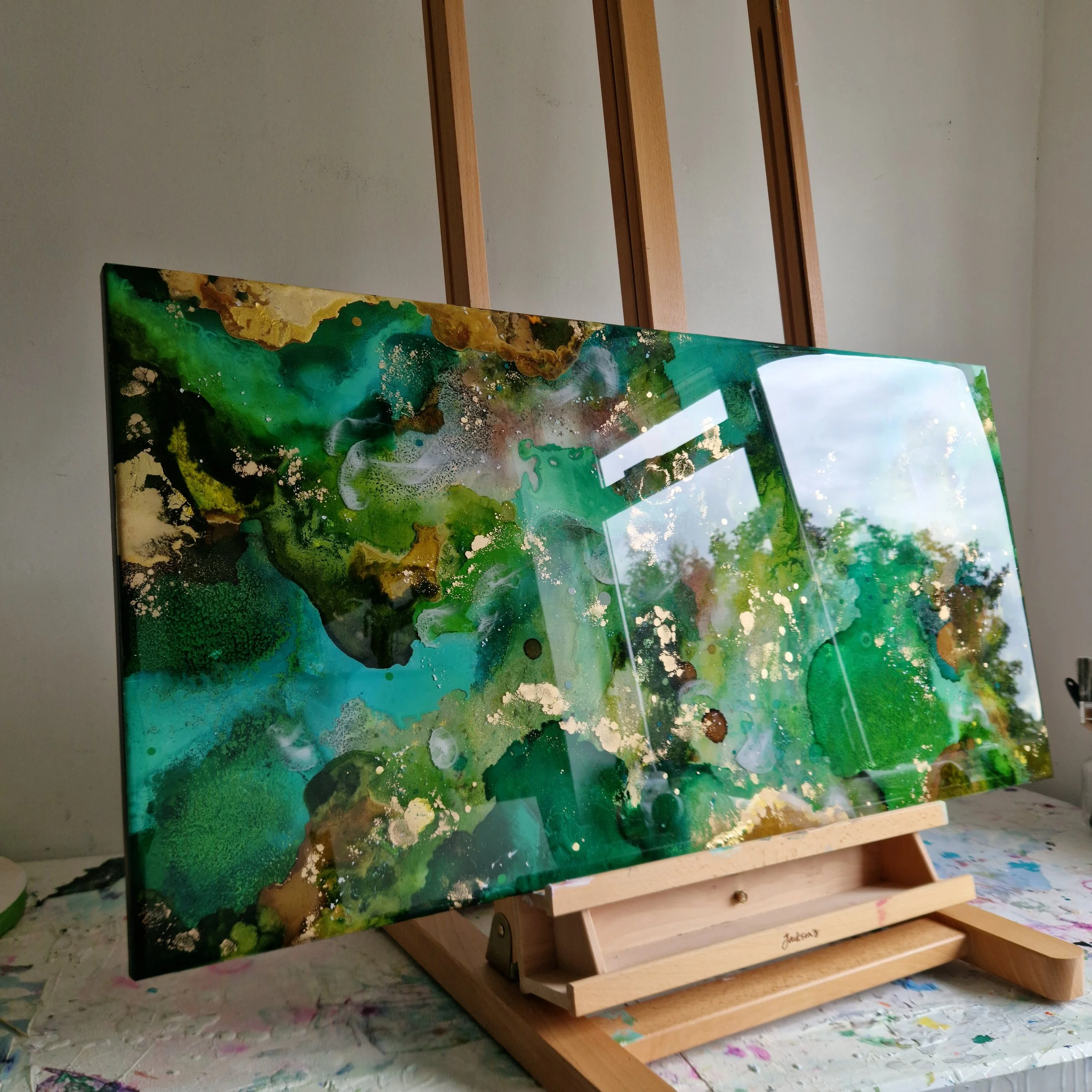 A forest inspired abstract painting with shades of green, black, gold, and white, displayed on an easel in an art studio. Original bespoke abstract resin ink and mixed media artwork created by artist Katie Alize, based in UK, Essex, London.
