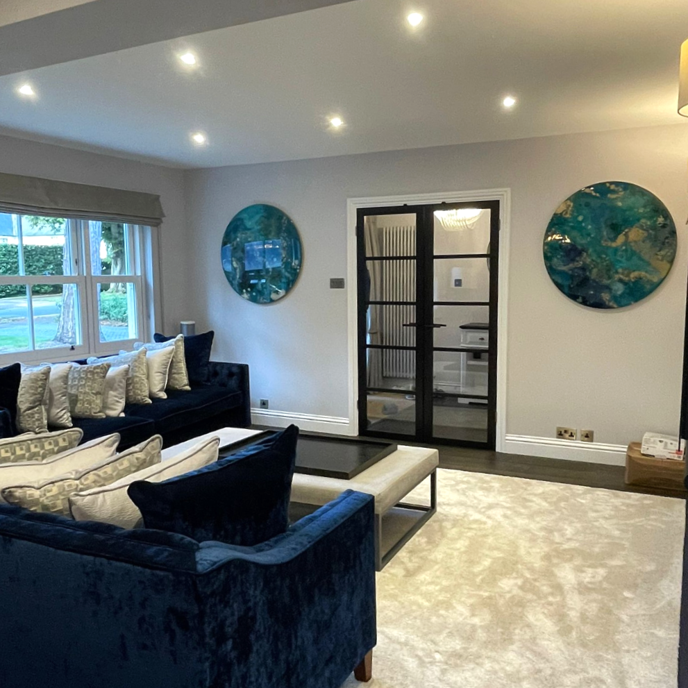 A modern living room with gray walls, a beige carpet, and a large window letting in natural light. There are navy blue sofas with multiple pillows, wall art with blue and teal resin mixed media abstract interior wall art designs made Katie Alize resi