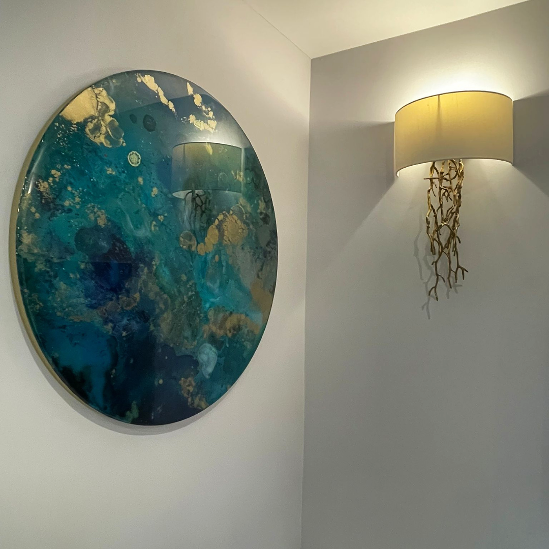 Round resin circle mixed media abstract interior large custom wall art designs made Katie Alize resin artist Essex London UK abstract wall art with blue, teal, and gold tones hanging on a white wall, and a wall sconce with a beige lampshade and decor