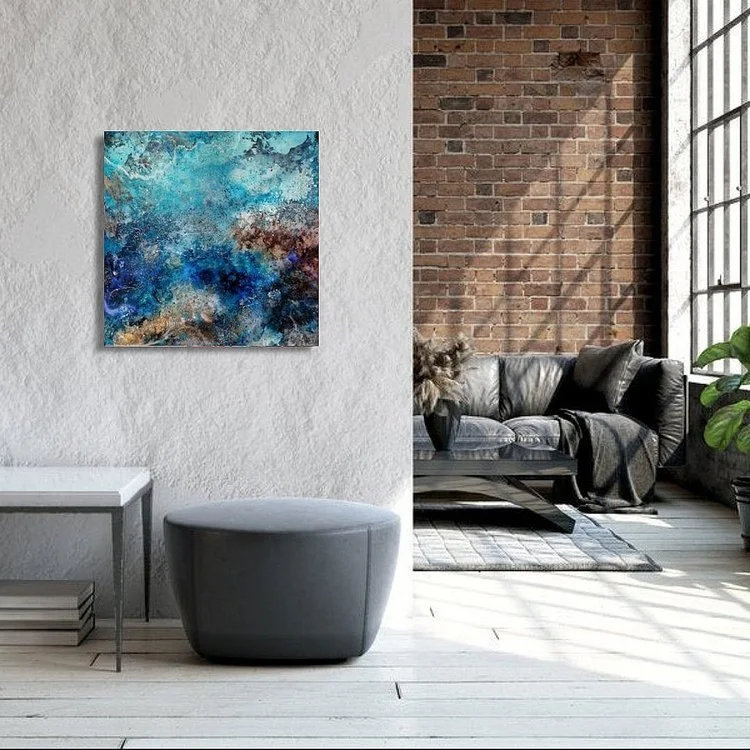 Interior living space featuring a white textured wall with abstract blue artwork, a gray upholstered ottoman, a white side table with stacked books, a black leather sofa on a patterned rug, a brick wall, large grid windows, and a potted plant.