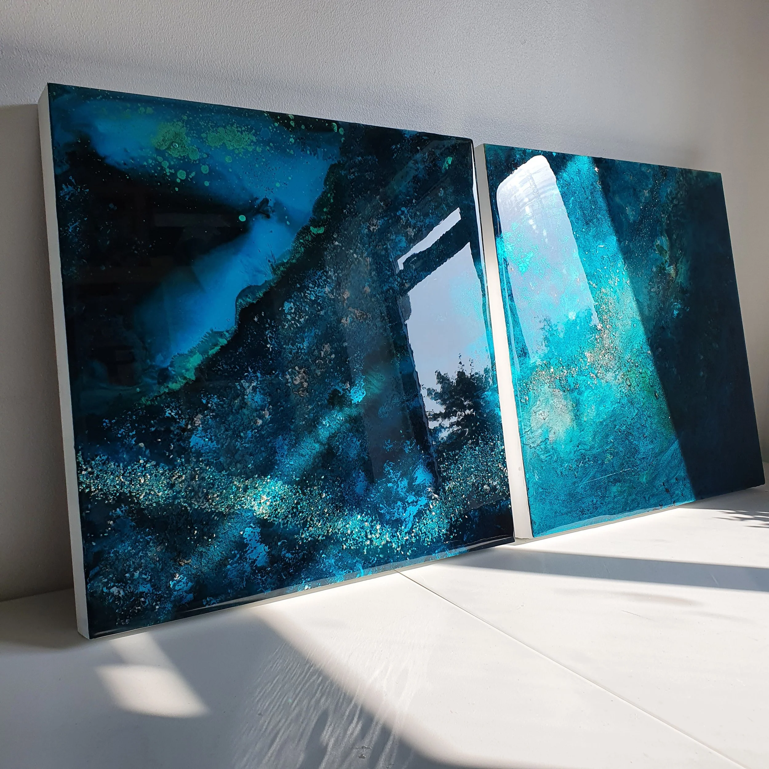 Two abstract blue and black resin ink mixed media paintings leaning against a white wall,  Abstract resin mixed media artwork created by UK based artist Katie Alize. Ocean resin painting, inspired by the sea. Blue resin art./