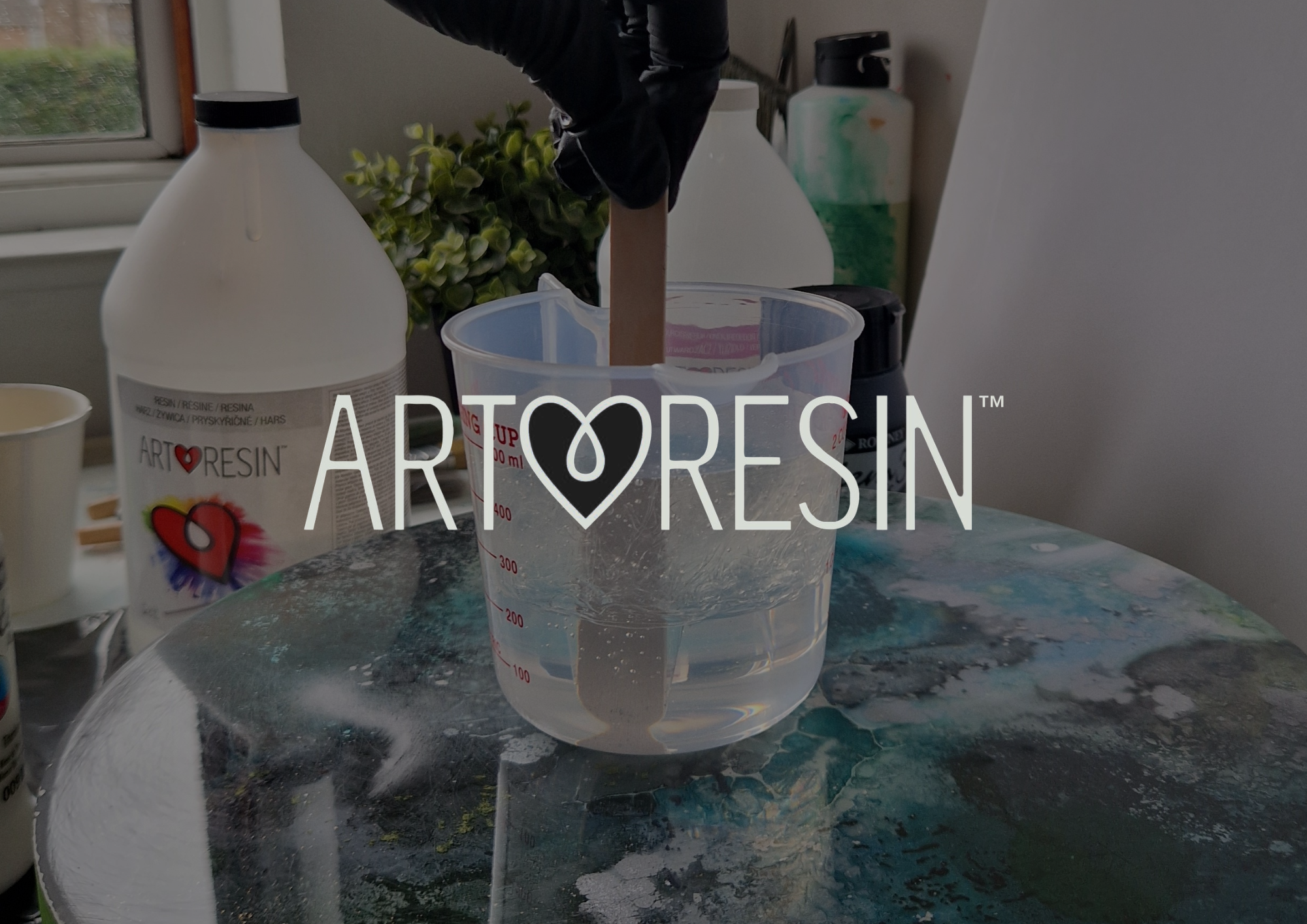 A plastic measuring cup on a table with art resin and a container of ArtResin brand resin. In the background, there are two bottles, a houseplant, and a window.
