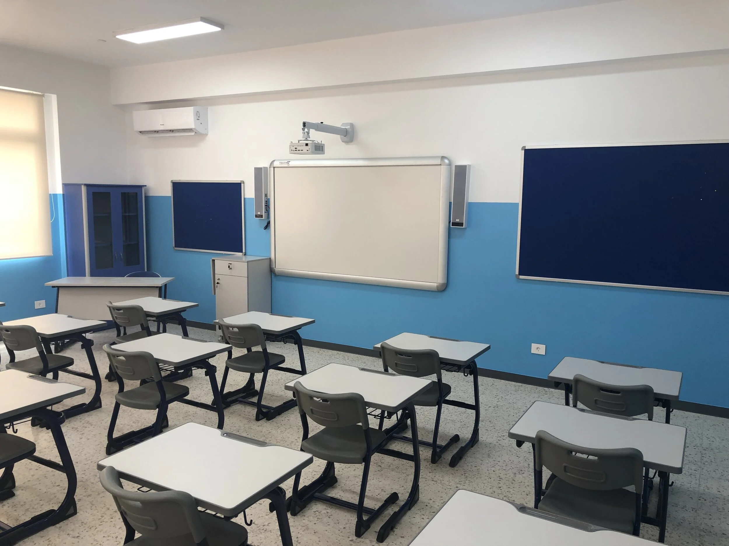 Empty classroom with blue and white walls, multiple gray student desks and chairs, a whiteboard, a chalkboard, digital screens, and an air conditioning unit.