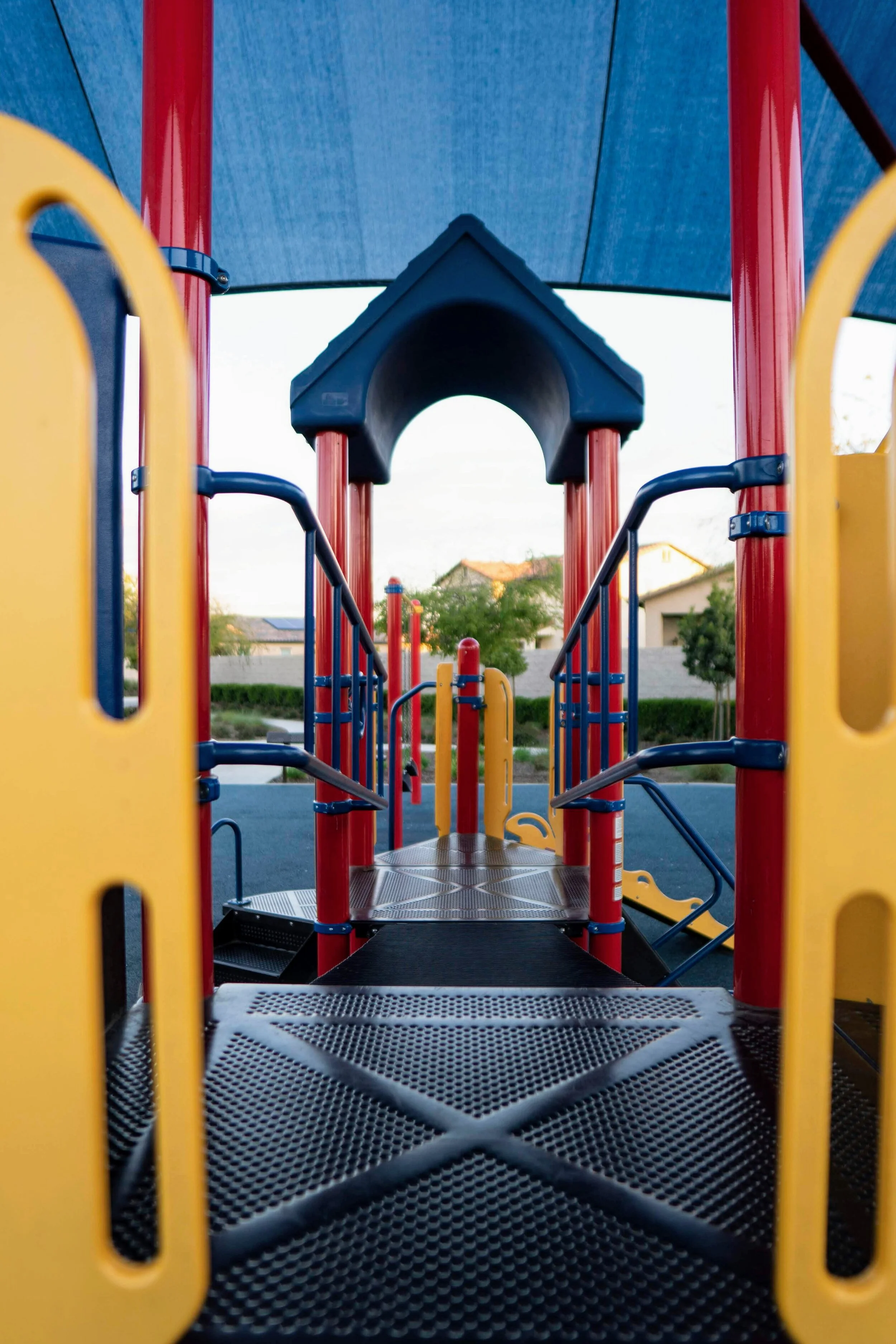 View of a colorful playground structure from the front, featuring a tunnel, red and yellow panels, and blue and red support poles, with houses and trees in the background.