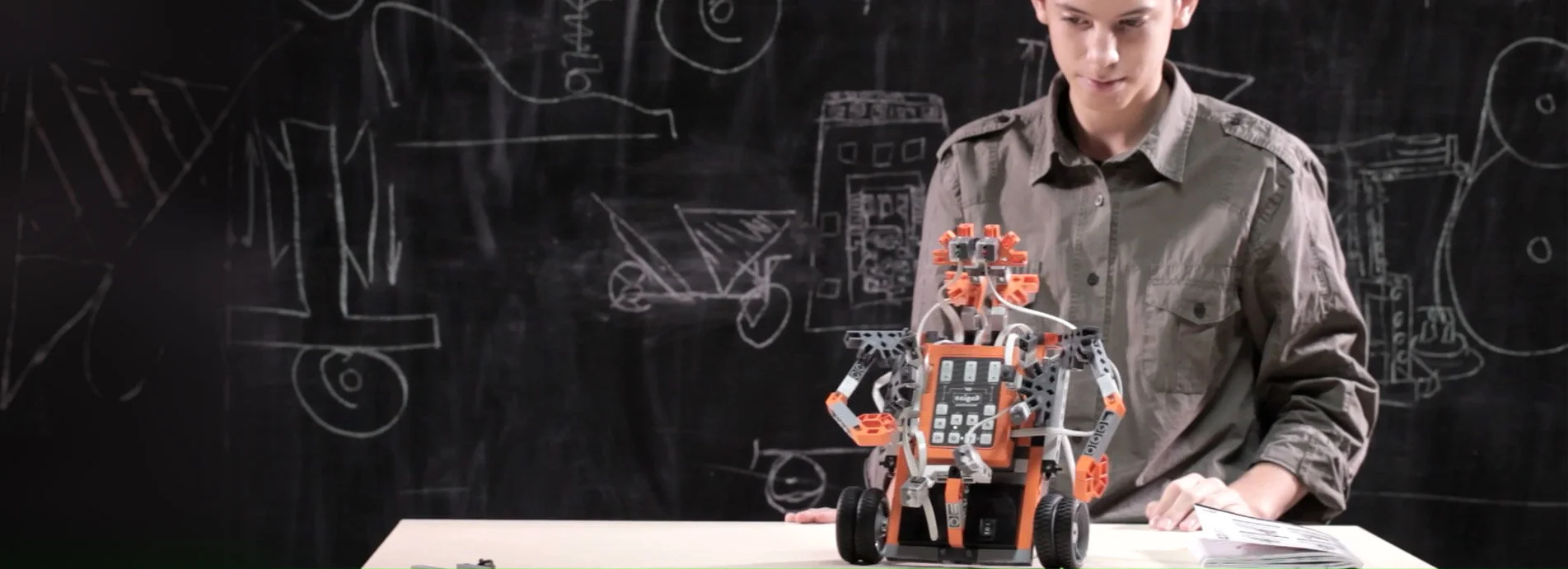 A young man in a gray shirt working on a robot made of orange and black parts on a table, with a blackboard filled with sketches and diagrams in the background.