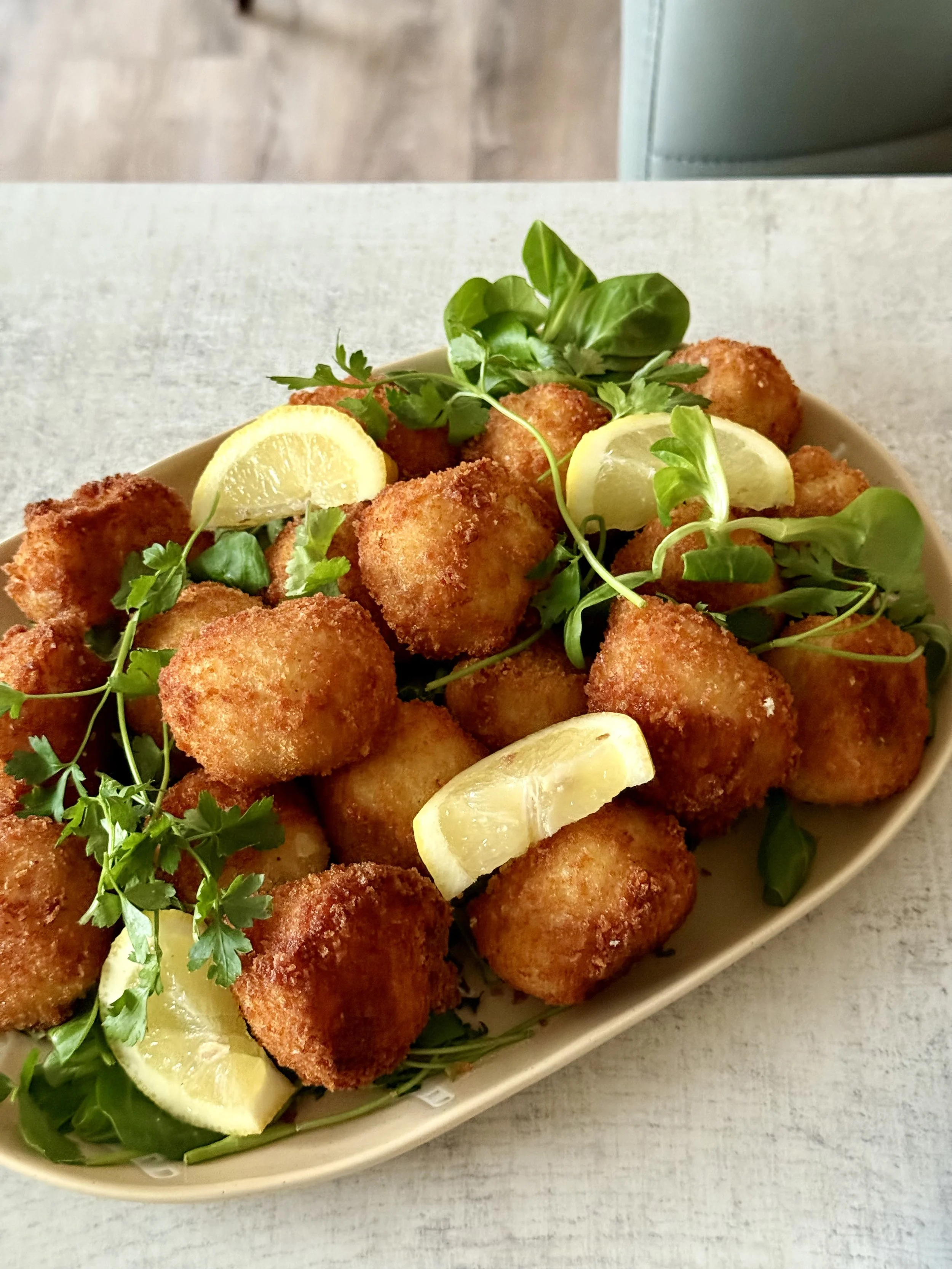 A white oval dish filled with breaded and fried fish balls garnished with fresh herbs and lemon wedges, placed on a light-colored surface.