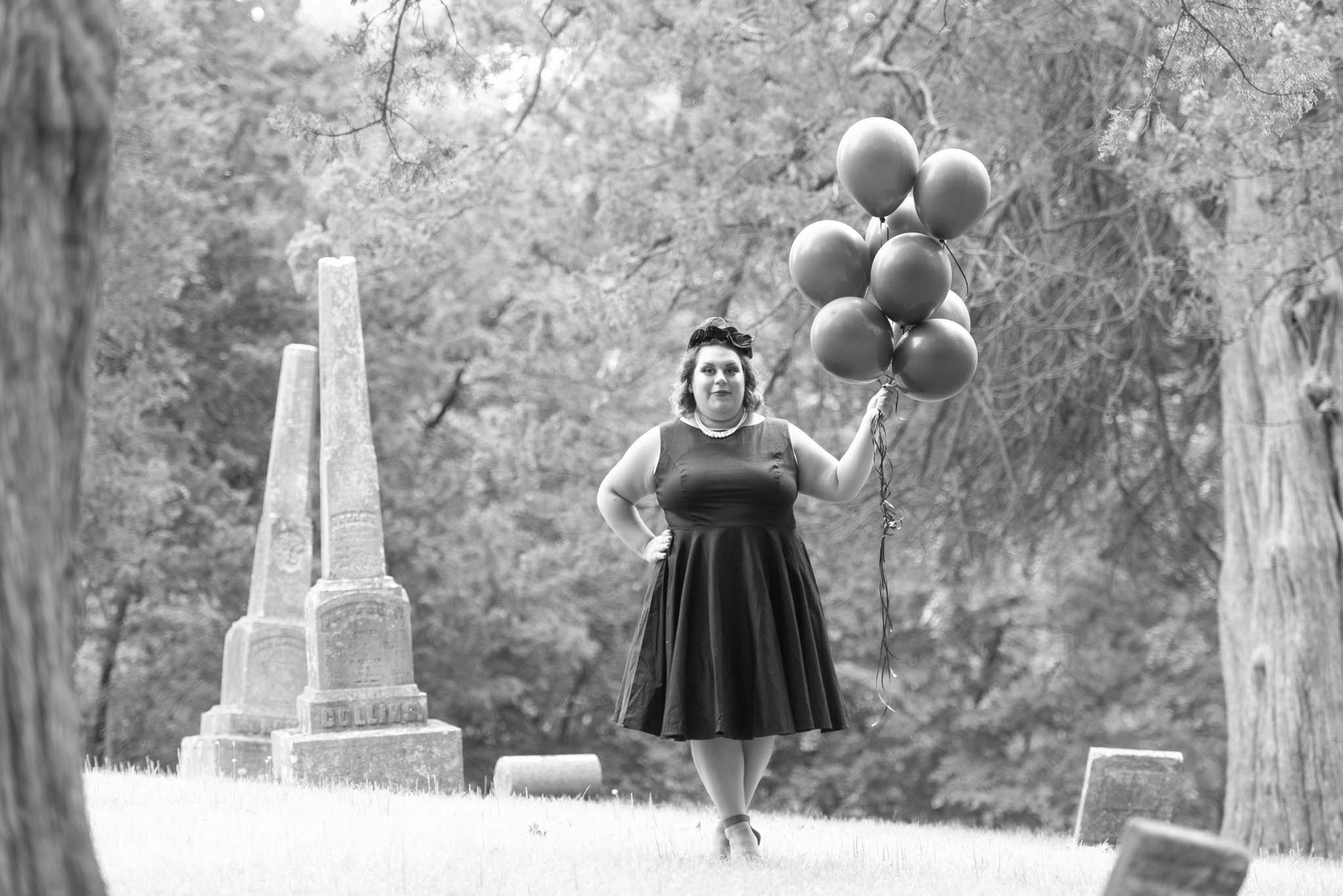  Samantha in black dress and vintage black hat poses in cemetery with black balloons. Photo is displayed in black and white. 