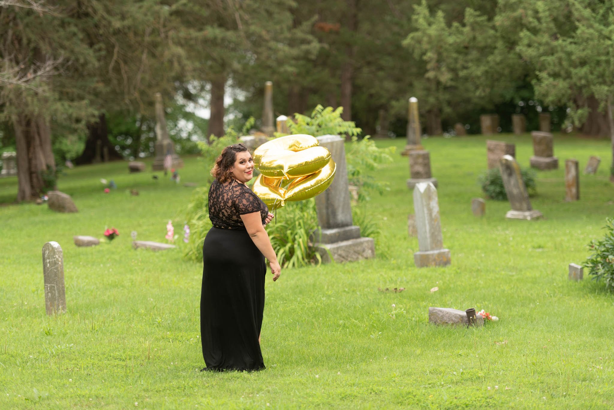  Samantha in black dress walks through cemetery carrying foil letter balloons and looks over her shoulder.  