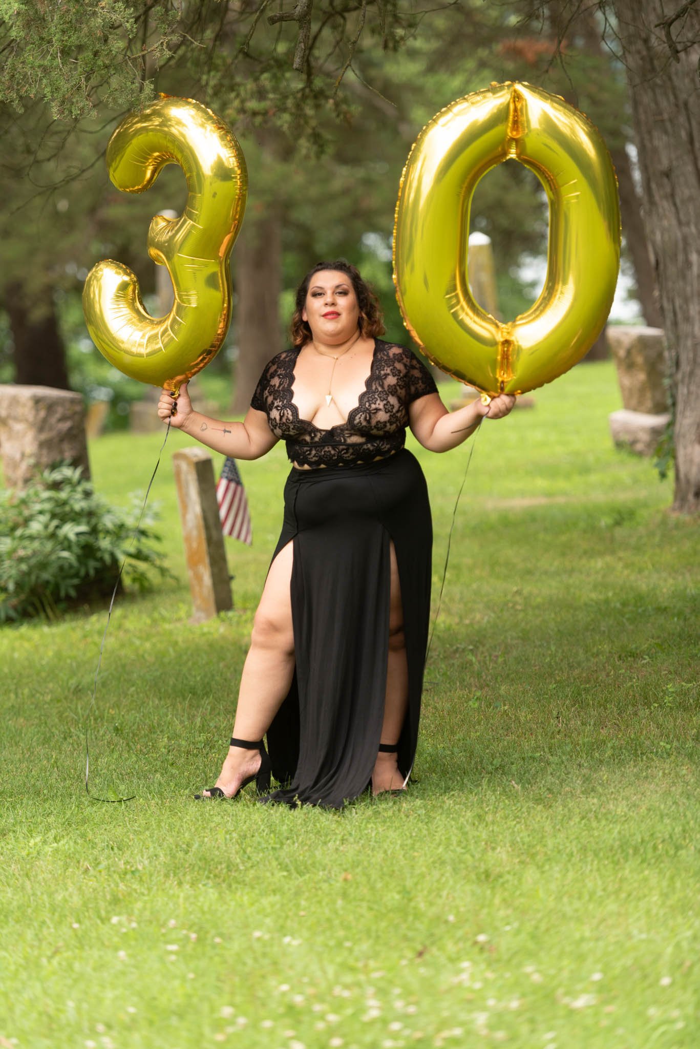  Samantha in black dress poses fiercely in cemetery holding gold foil balloons displaying the number 30. 