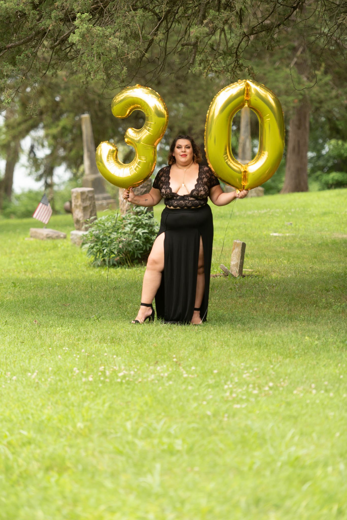  Samantha in black dress poses fiercely in cemetery holding gold foil balloons displaying the number 30. 