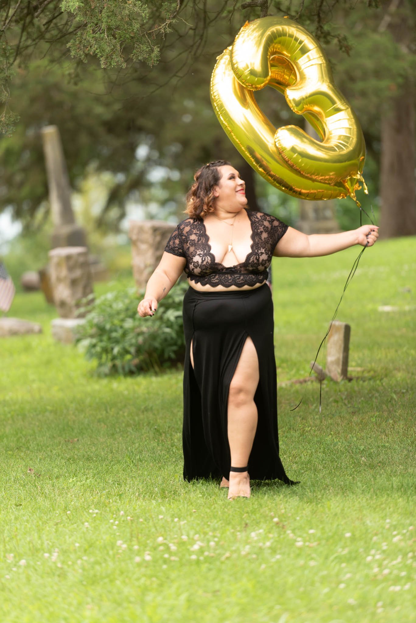  Samantha in black dress walks through cemetery carrying foil letter balloons and looks over her shoulder.  