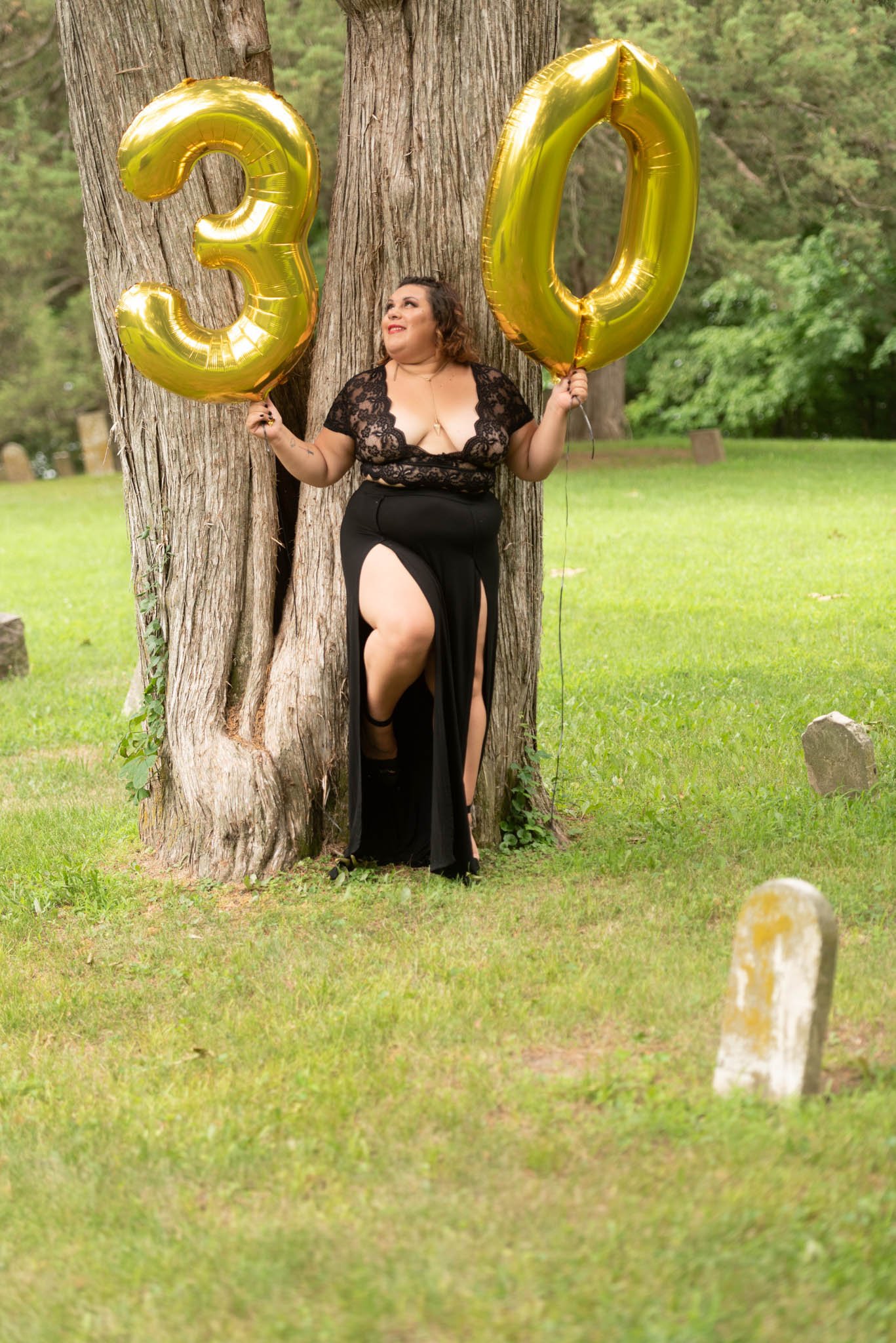  Samantha in black dress poses fiercely against a tree in cemetery holding gold foil balloons displaying the number 30. 