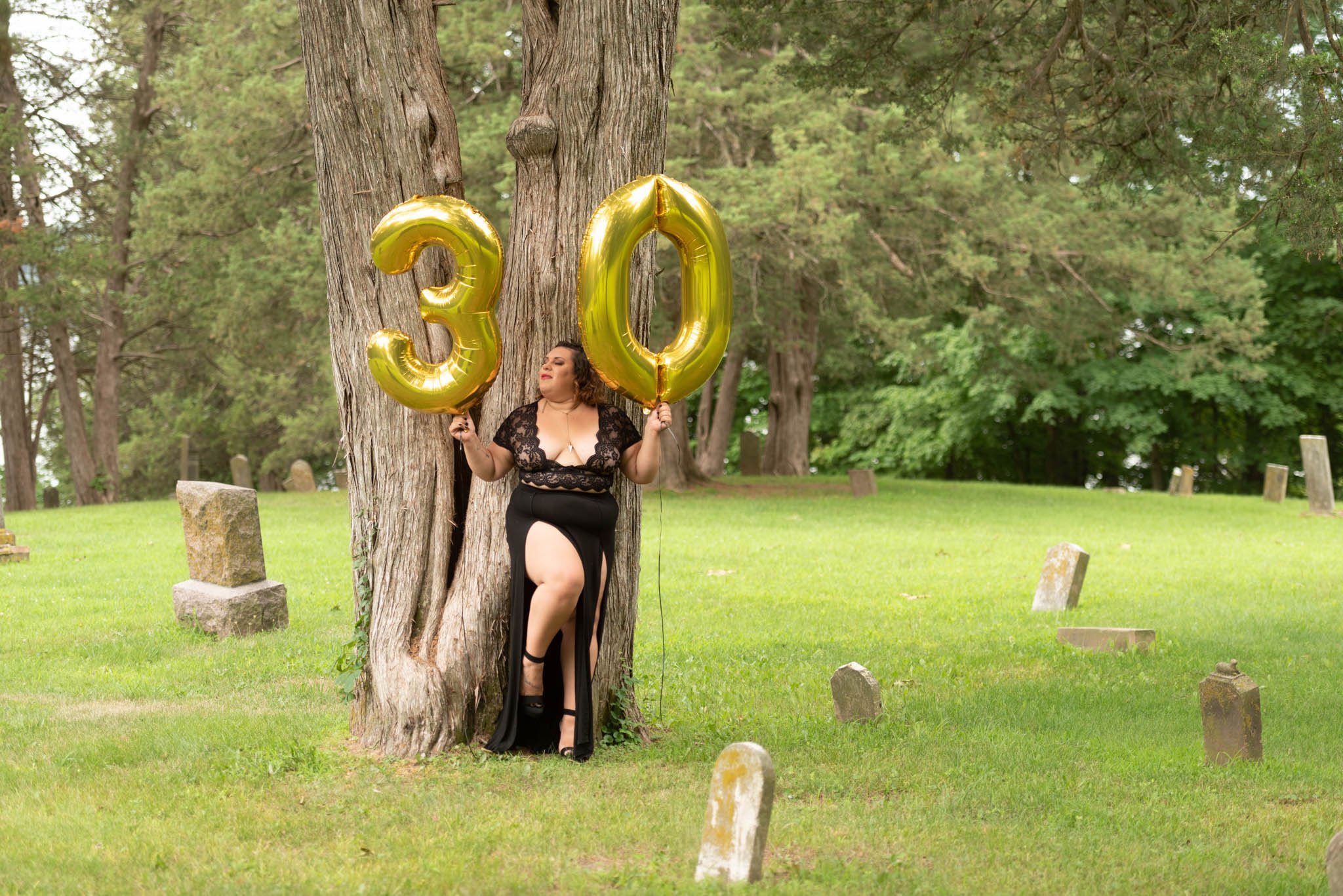  Samantha in black dress poses fiercely against a tree in cemetery holding gold foil balloons displaying the number 30. 