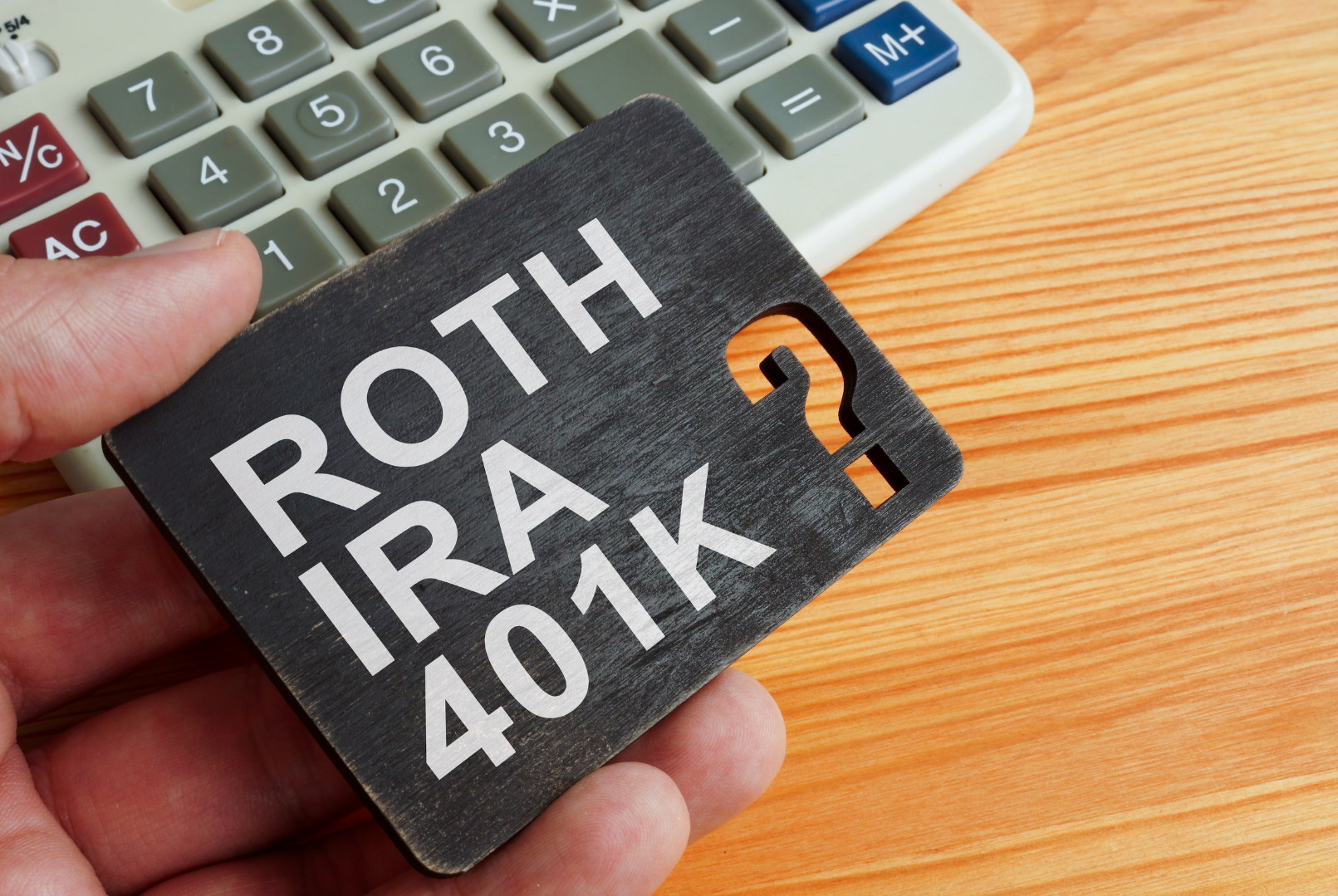 Close-up of a hand holding a card with 'Roth IRA 401K' written on it, with a question mark cut-out, placed on a wooden desk next to a calculator
