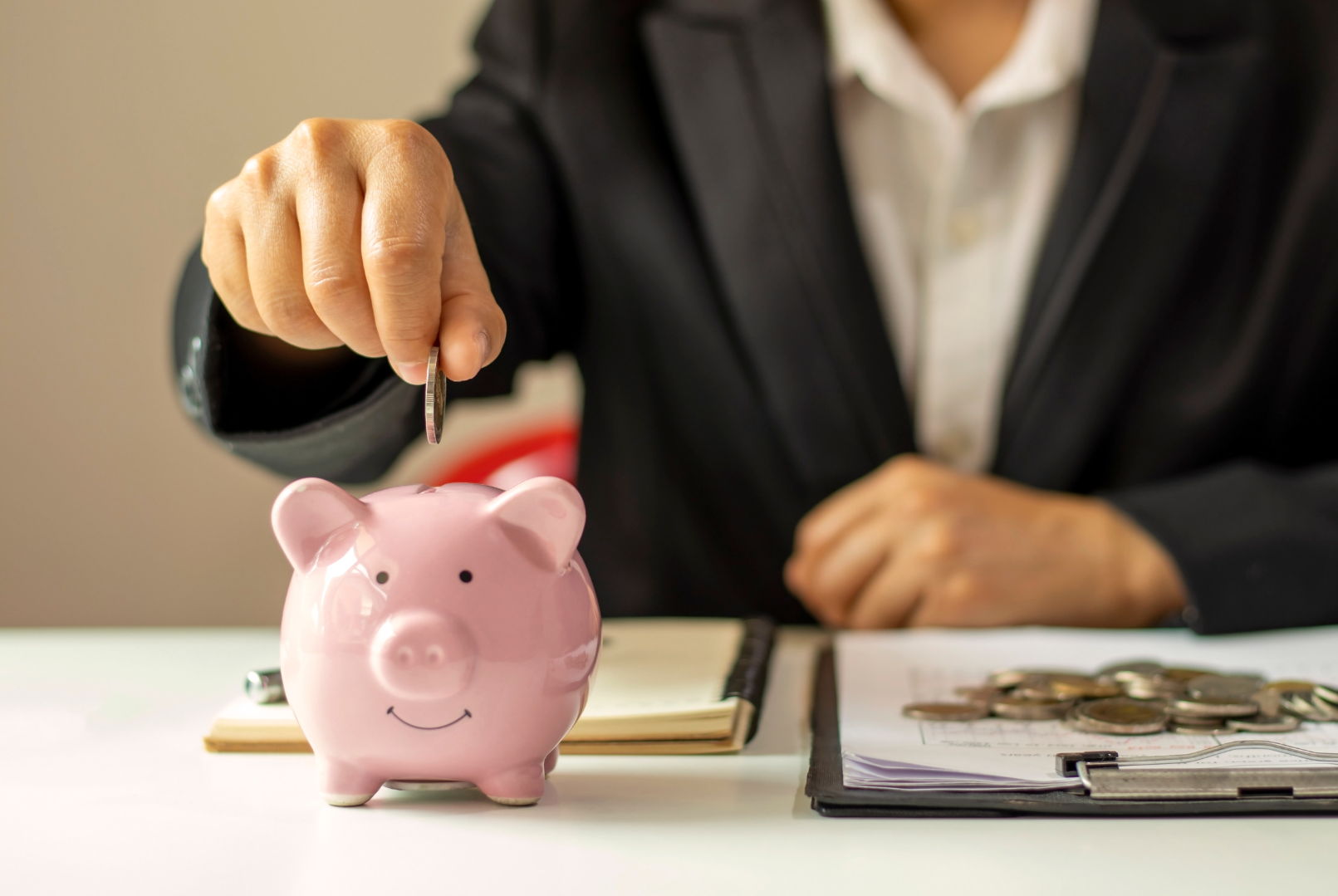 A financial advisor placing a coin into a pink piggy bank, with a notebook and keys on the desk in front of them.