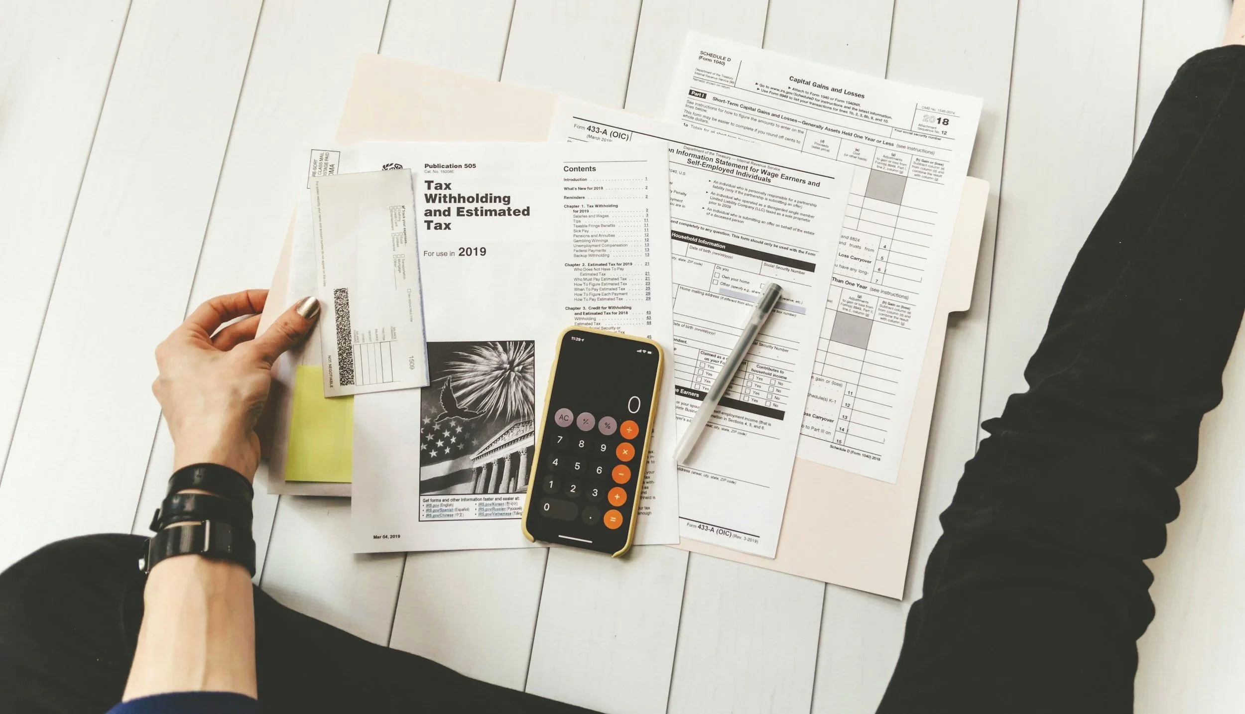 Overhead view of tax-related documents spread on a table, envelopes, and a smartphone calculator showing numbers, suggesting someone is preparing or reviewing taxes.