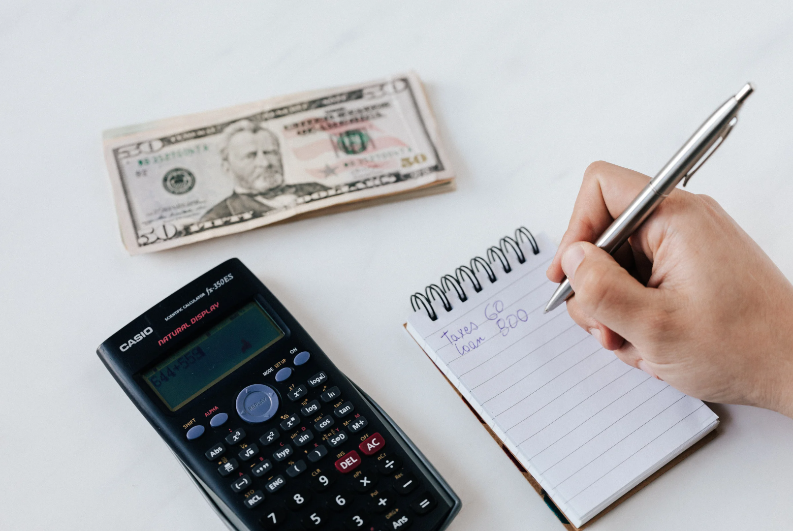 Financial milestones: A person calculating expenses and writing down notes, with a calculator, $50 bill, and a notepad displaying 'Taxes 60' and 'Loan 800.