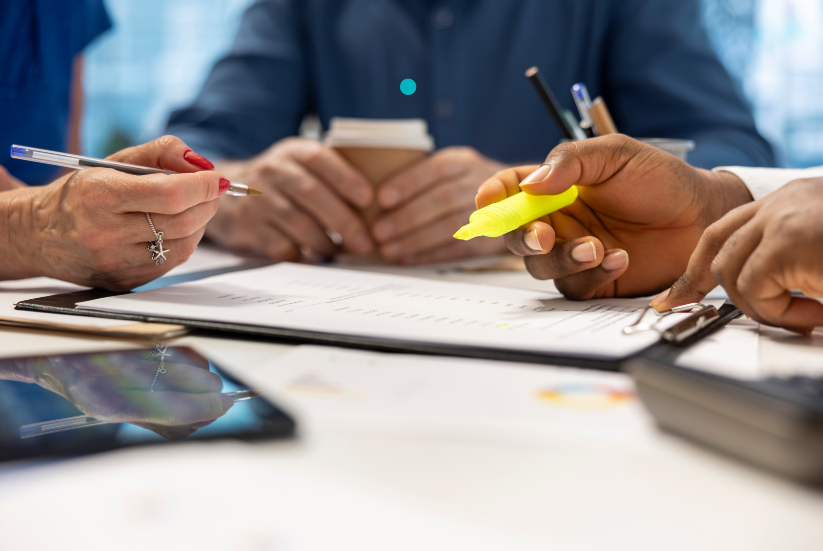 Hands of several people in a business meeting reviewing a document on a table, with one person holding a yellow highlighter and others holding pens and a coffee cup, suggesting collaborative discussion and document editing.