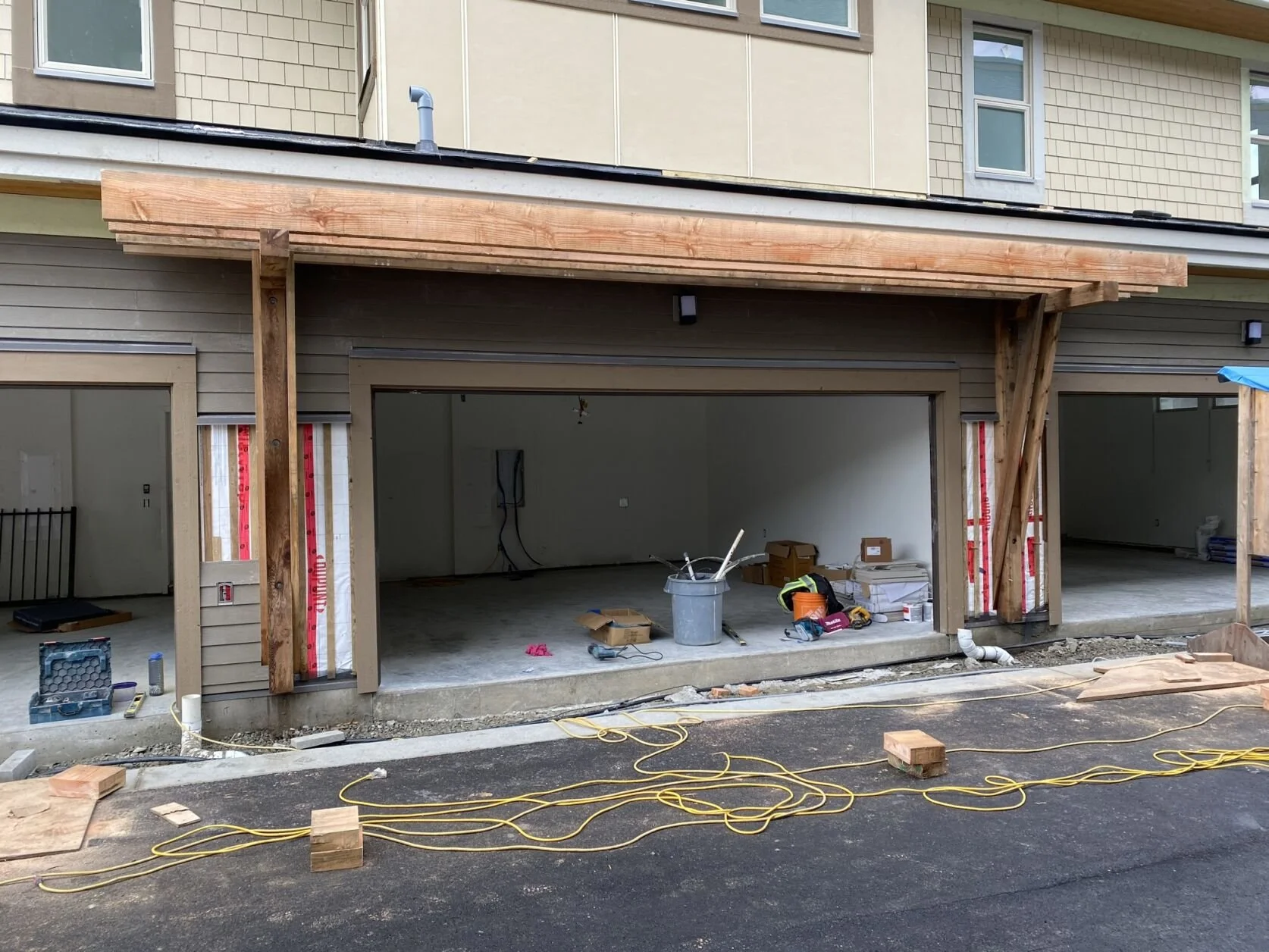 Construction site of a building with two garage doors, unfinished exterior with wooden framing, and construction tools and materials on the ground.