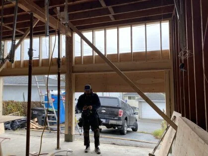Construction site showing a partially built wooden structure, a man in dark work clothes and helmet working, construction tools nearby, and a residential area in the background.
