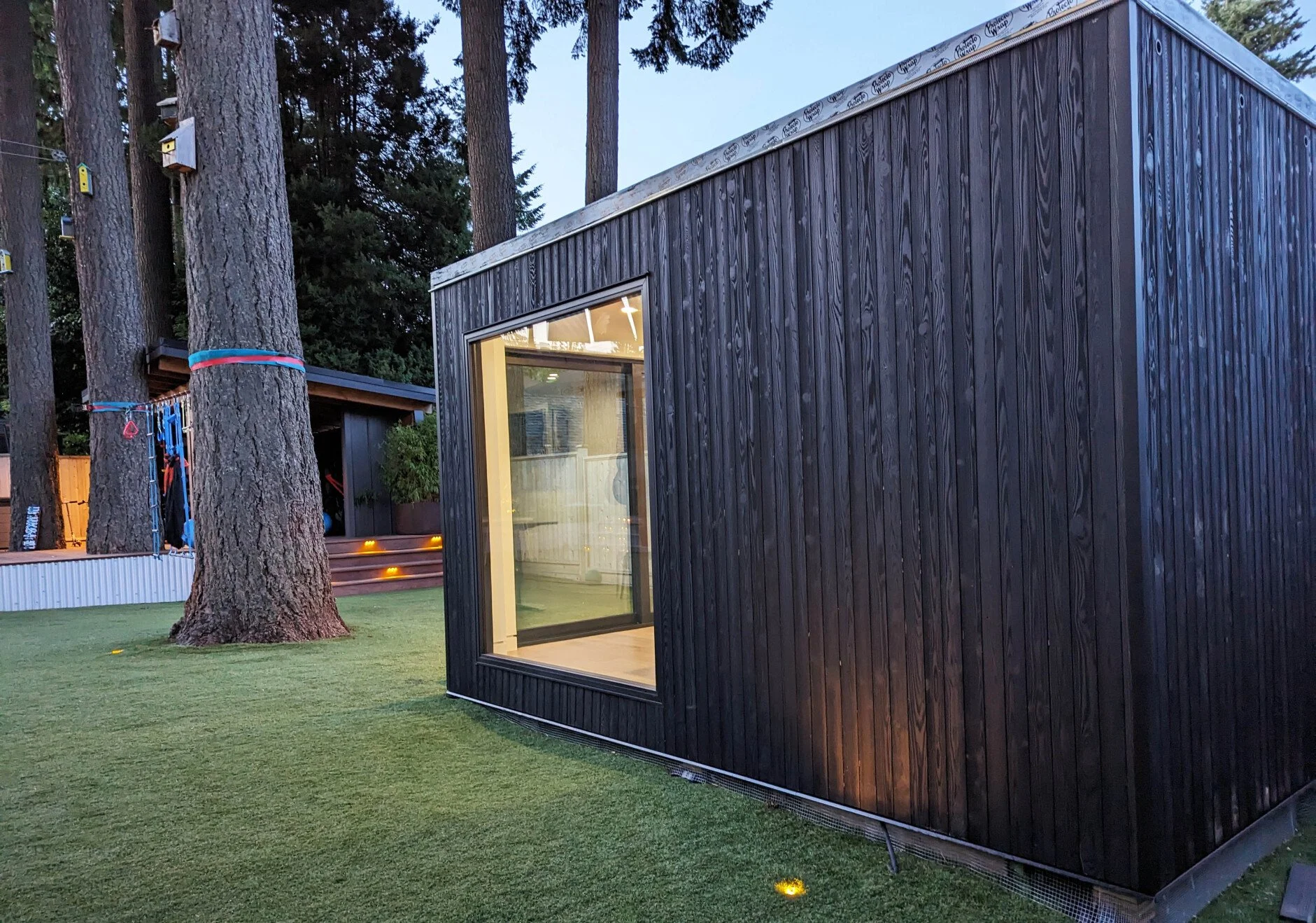 A small modern black wooden building with a large glass window, situated on a grassy area surrounded by tall trees at dusk.