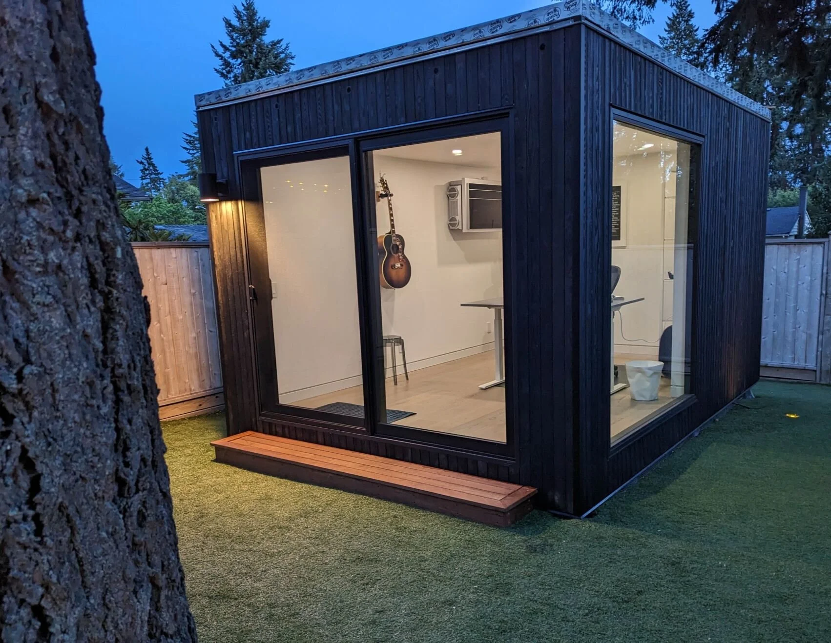 A tiny modern black wooden studio with large glass windows on three sides, situated in a yard with artificial grass. Inside, a guitar hanging on the wall, a small TV, a desk, and an office chair are visible.