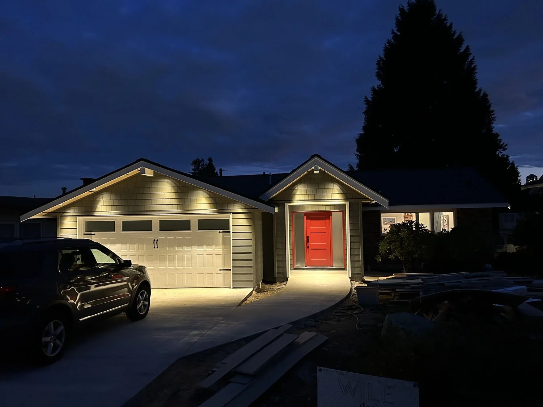 A house at night illuminated by exterior lights, with a red front door, a garage, a parked car, and dark clouds in the sky.