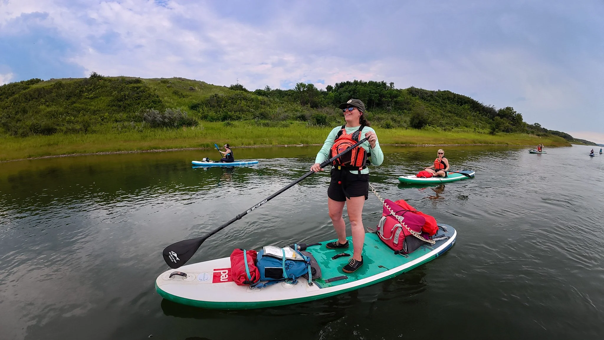 Group of women paddleboarding on a river with grassy hills in the background, Overnight Stand-up Paddleboard Camping Adventure
