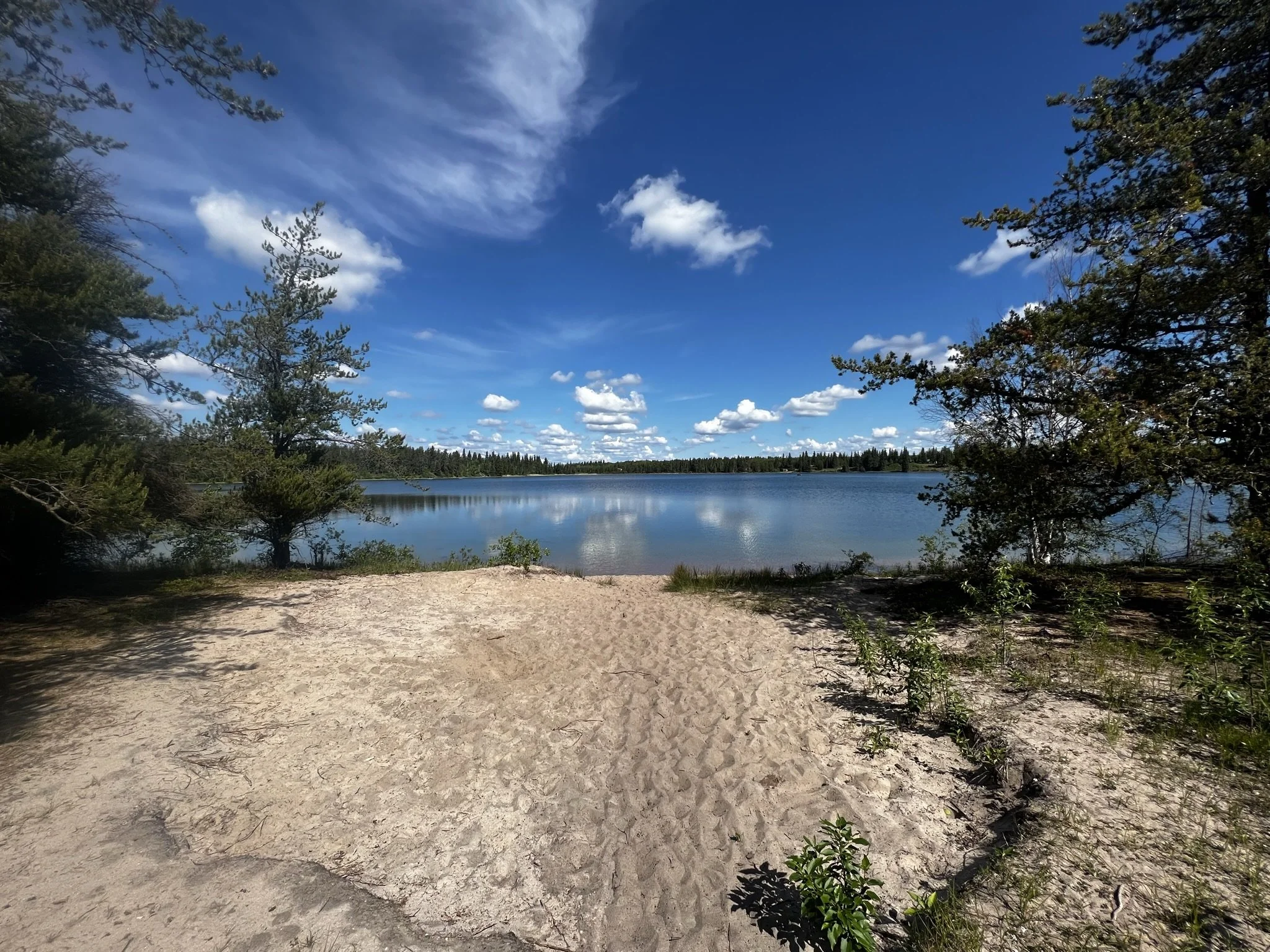 A sandy beach shoreline overlooking a calm lake with trees on both sides, under a blue sky with scattered clouds.
